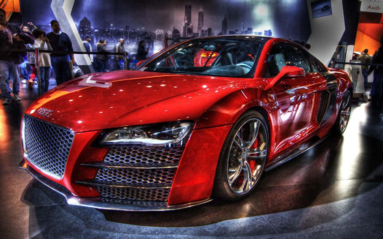 奧迪 桌布 壁紙 — 一輛光澤的紅色奧迪R8跑車在車展上，在明亮的燈光下閃閃發光，背景是城市天際線。