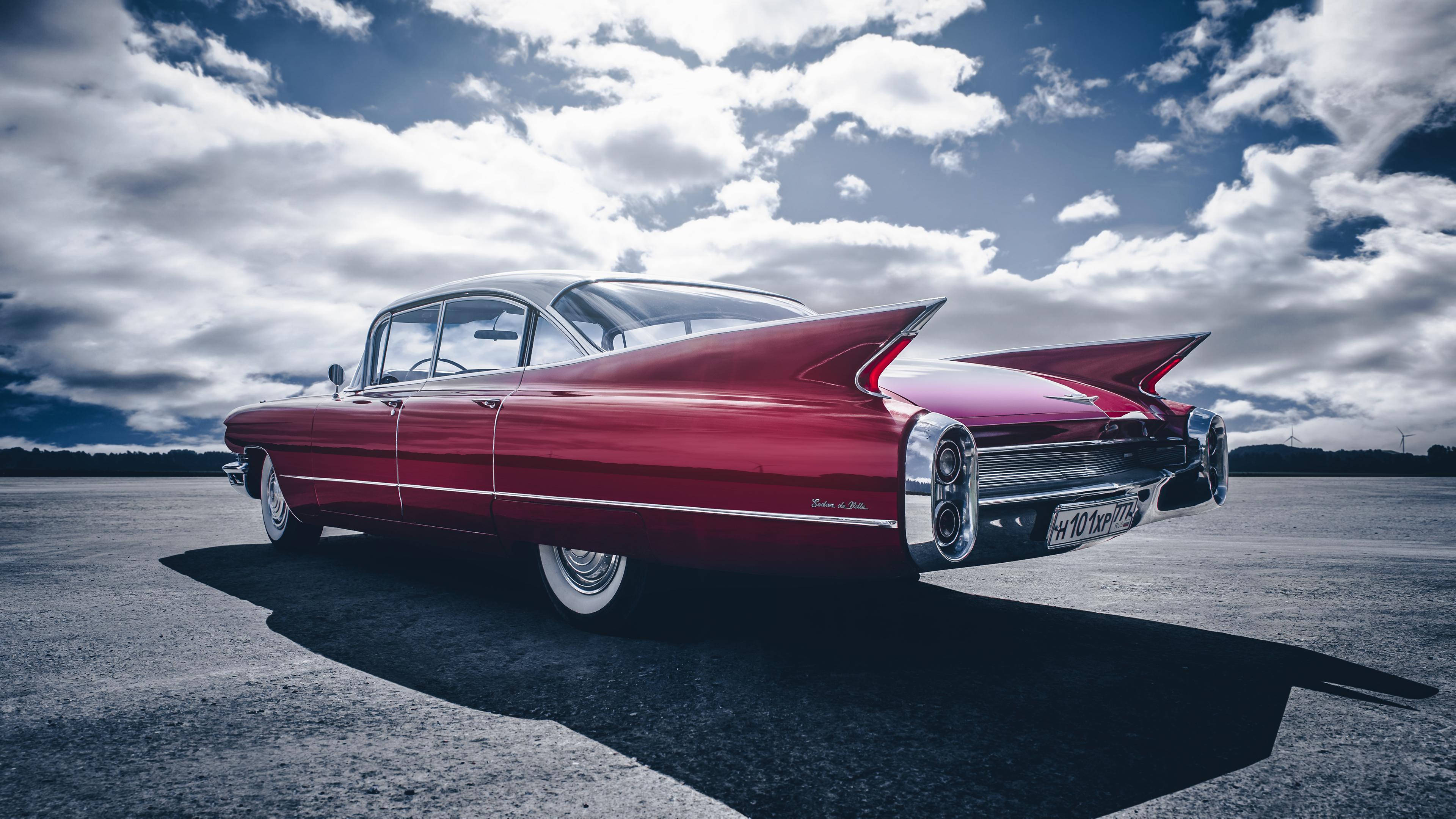 Cadillac 桌布 壁紙 — 一輛光澤的紅色復古凱迪拉克，帶有戲劇性的尾鰭，獨自停在開闊的瀝青場地上，天空多雲而戲劇化。