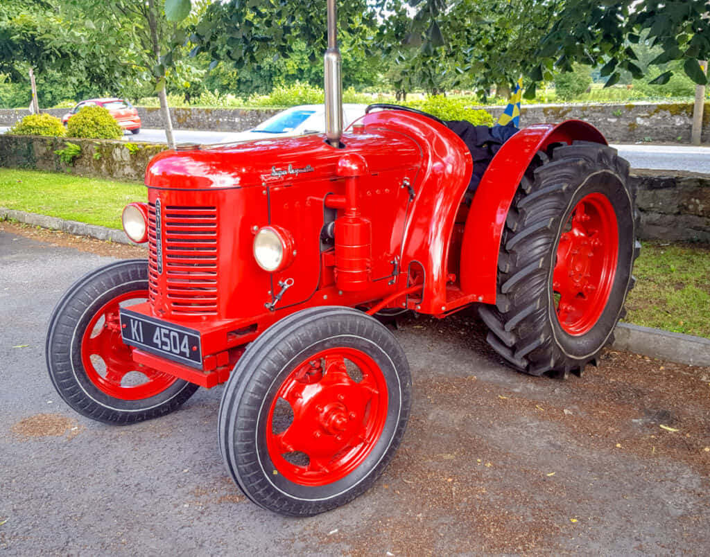 Tractor 桌布 壁紙 — 一輛光澤的紅色復古拖拉機，黑色輪胎停在瀝青路上，旁邊有綠草和樹木。