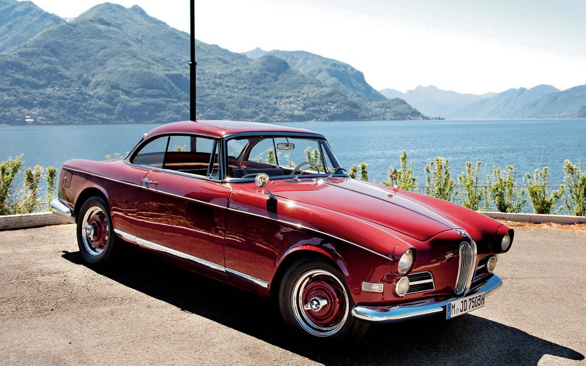 classic bmw 桌布 壁紙 — 一輛光澤的紅色復古跑車停泊在寧靜的湖邊，背景是山巒，在明亮的日光下。