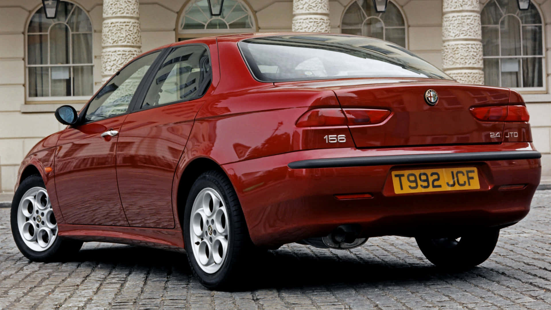 Alfa Romeo 156 桌布 壁紙 — 一輛光澤的紅色愛快羅密歐156轎車停在古典建築前的鵝卵石街道上。