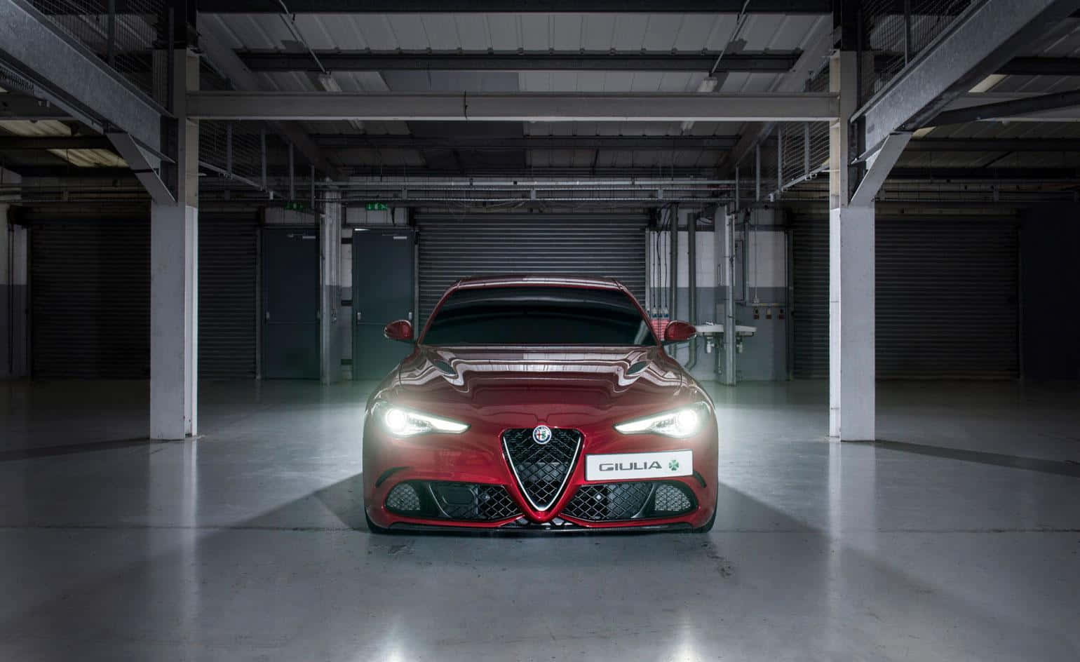 Alfa Romeo Giulia 桌布 壁紙 — 一輛光澤的紅色愛快羅密歐Giulia跑車在明亮的車頭燈下，於空曠的工業車庫中閃耀。