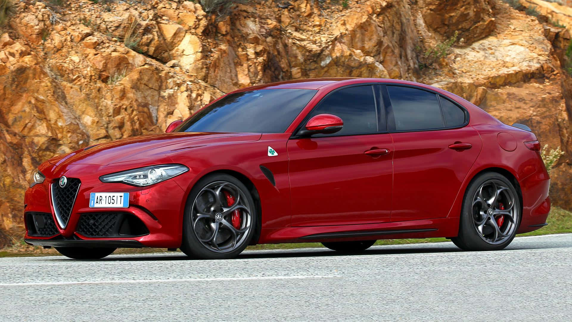 Alfa Romeo Giulia 桌布 壁紙 — 一輛光澤的紅色愛快羅密歐轎車，配備黑色輪圈和紅色制動卡鉗，停在鋪砌的道路上，旁邊是崎嶇的橙色岩壁。
