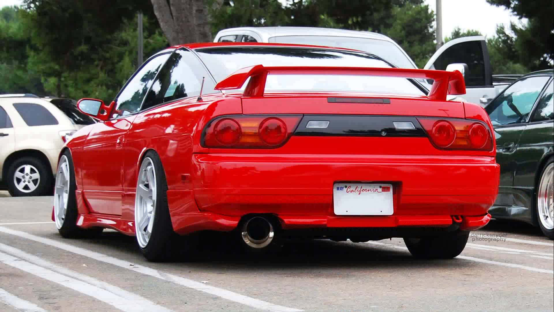 Nissan 180sx 桌布 壁紙 — 一輛光澤的紅色日產Skyline R34跑車停放在停車場，背景有樹木和其他車輛。