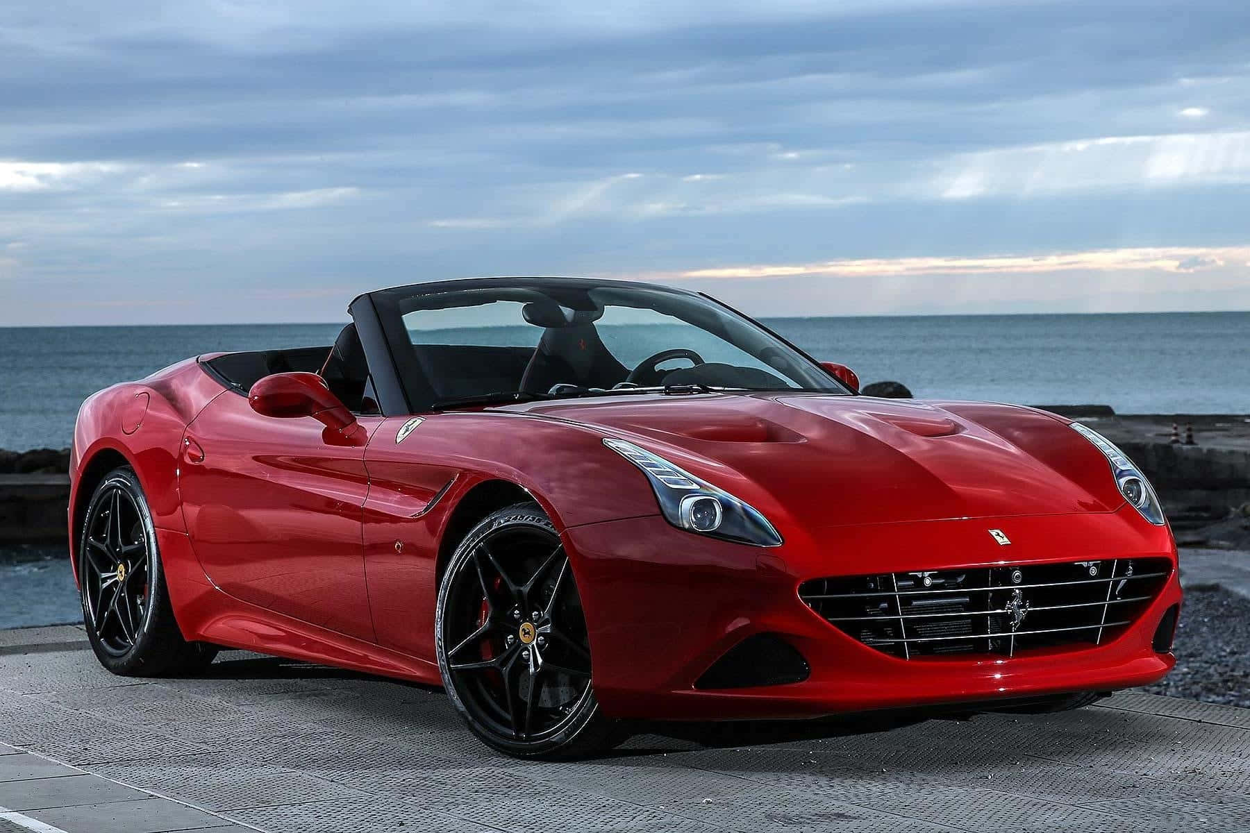 Ferrari California T 桌布 壁紙 — 一輛光澤的紅色法拉利敞篷車停泊在石碼頭上，背景是海洋和多雲的天空。