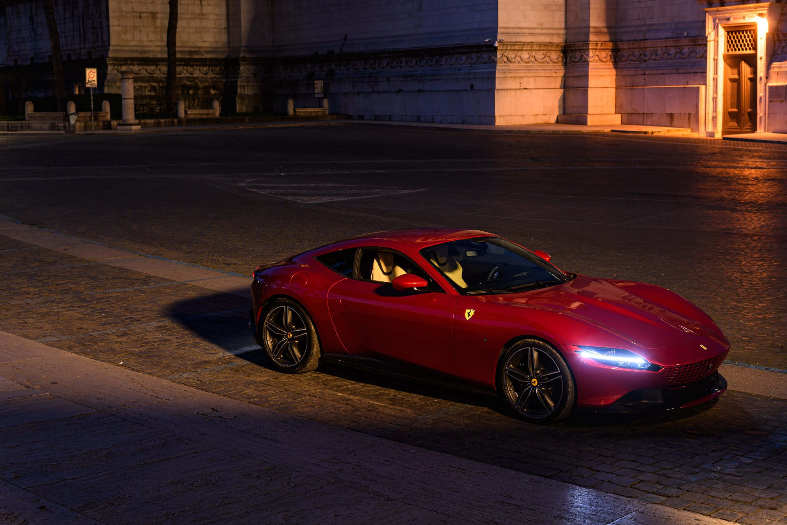 Ferrari Roma 桌布 壁紙 — 一輛光澤的紅色法拉利跑車在夜間停放在潮濕的鵝卵石街道上，在溫暖的路燈下閃耀。