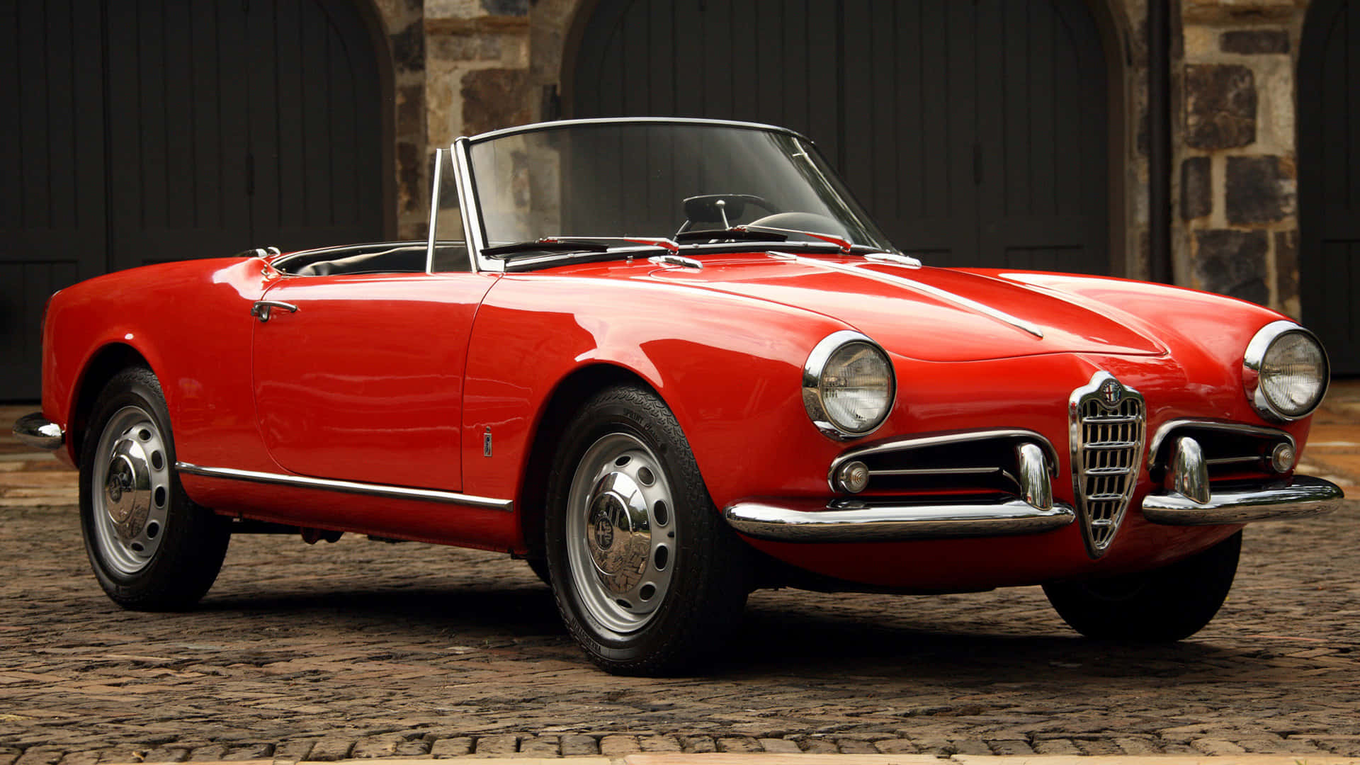 Alfa Romeo Spider 桌布 壁紙 — 一輛光澤的紅色經典愛快羅密歐Spider敞篷車停在鵝卵石街道上，背景有石拱門。