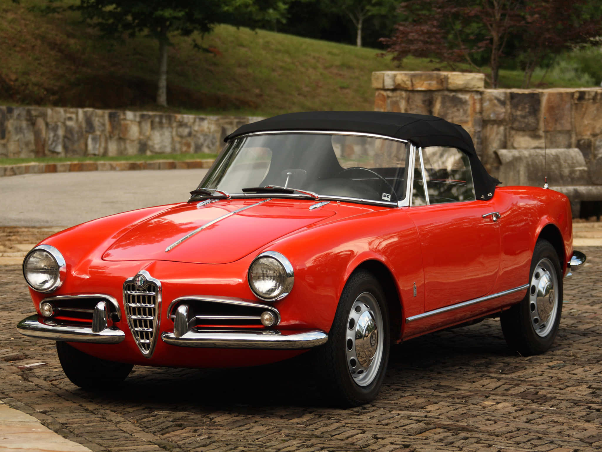Alfa Romeo Spider 桌布 壁紙 — 一輛光澤的紅色經典愛快羅密歐Spider敞篷車，配黑色軟頂，停放在石牆和綠植旁的鵝卵石車道上。