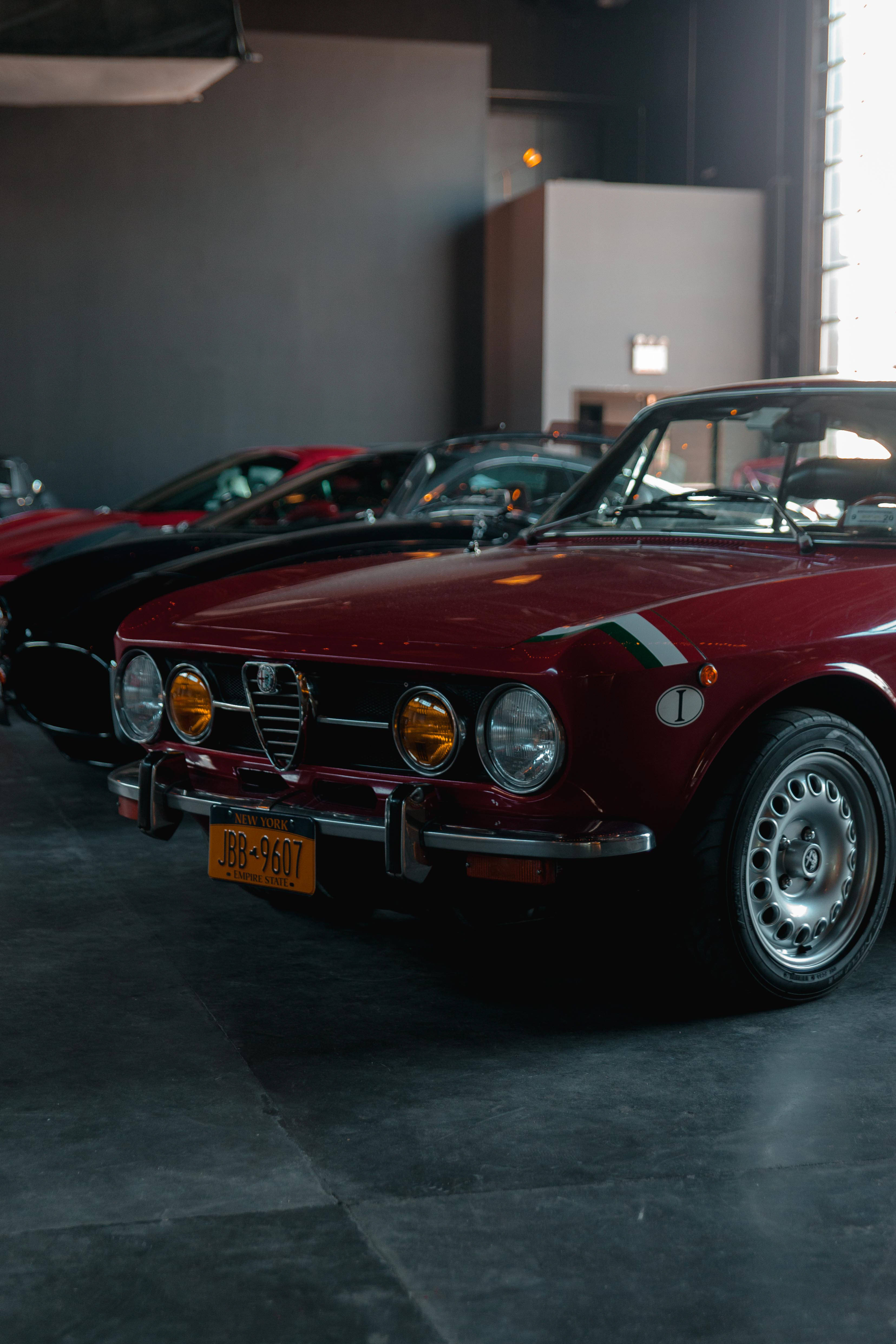 Alfa Romeo 桌布 壁紙 — 一輛光澤的紅色經典愛快羅密歐Spider在柔和光線下閃耀，背景是時尚現代汽車展廳中的其他復古汽車。