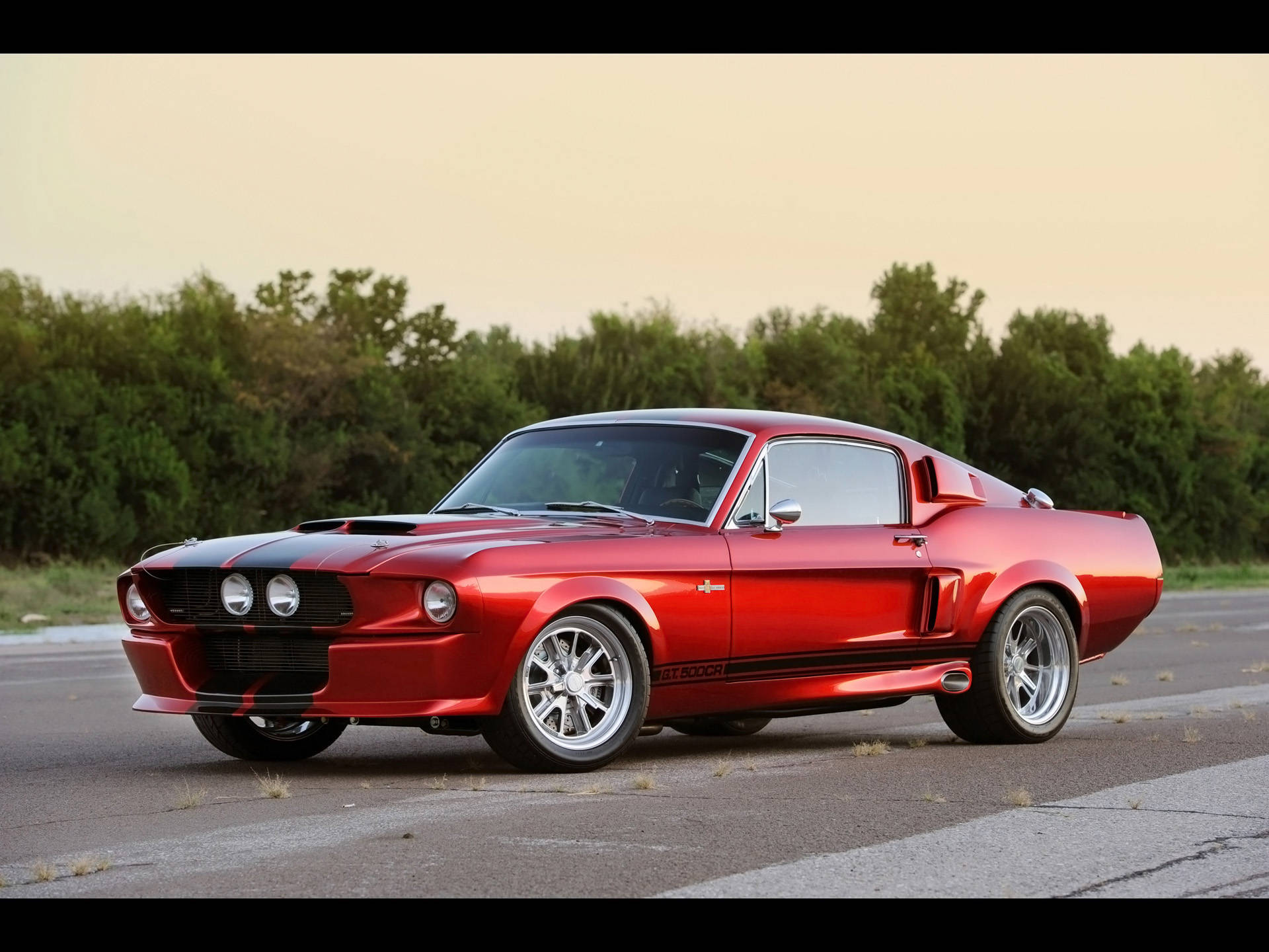 shelby iPhone 桌布 壁紙 — 一輛光澤的紅色經典肌肉車，帶有黑色賽車條紋，在黃金時段停在瀝青路上。
