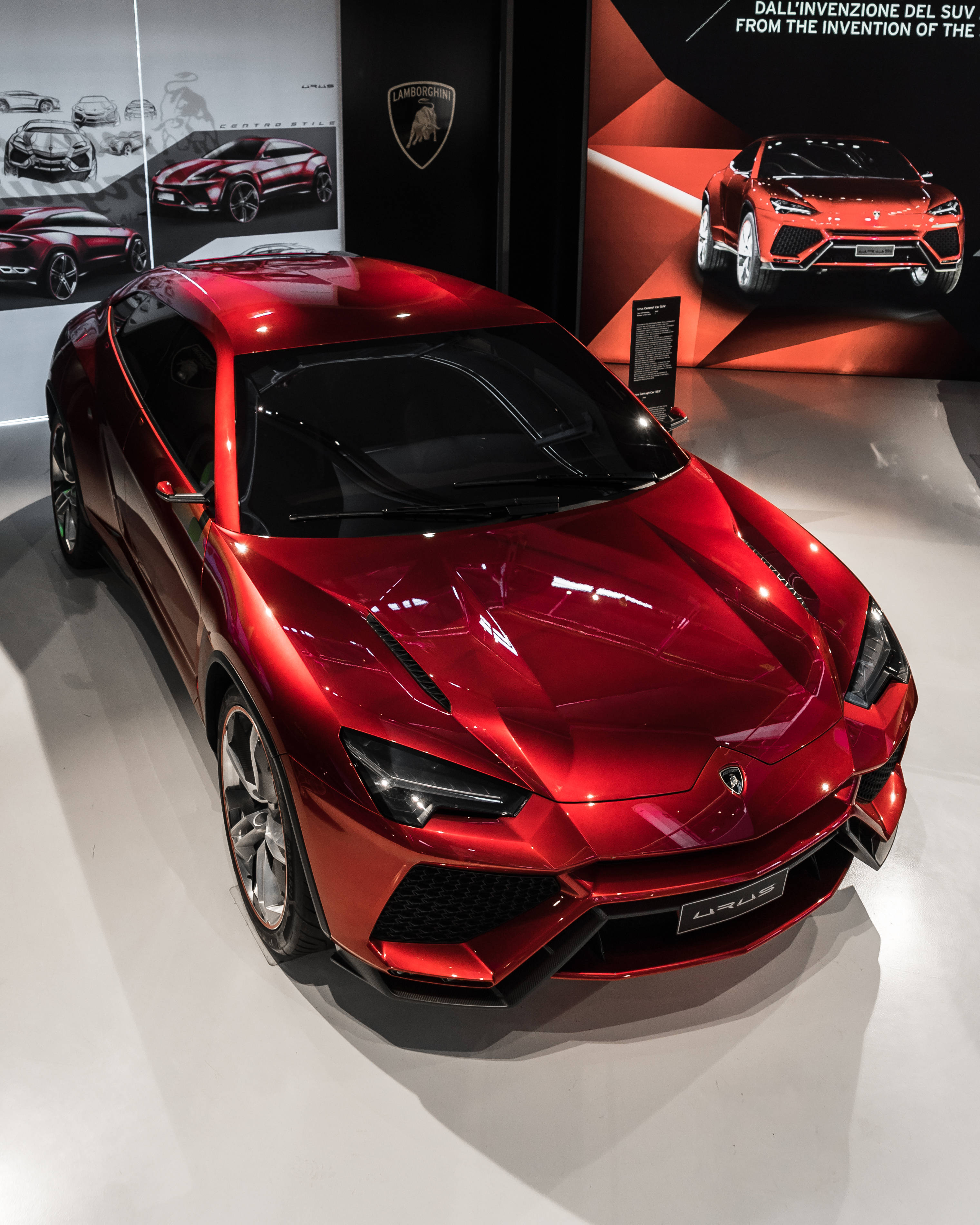 Luxury 桌布 壁紙 — 一輛光澤的紅色藍寶堅尼 Urus SUV 在明亮燈光下，於一個時尚現代、帶有品牌牆壁和概念藝術的展示間中閃閃發光。