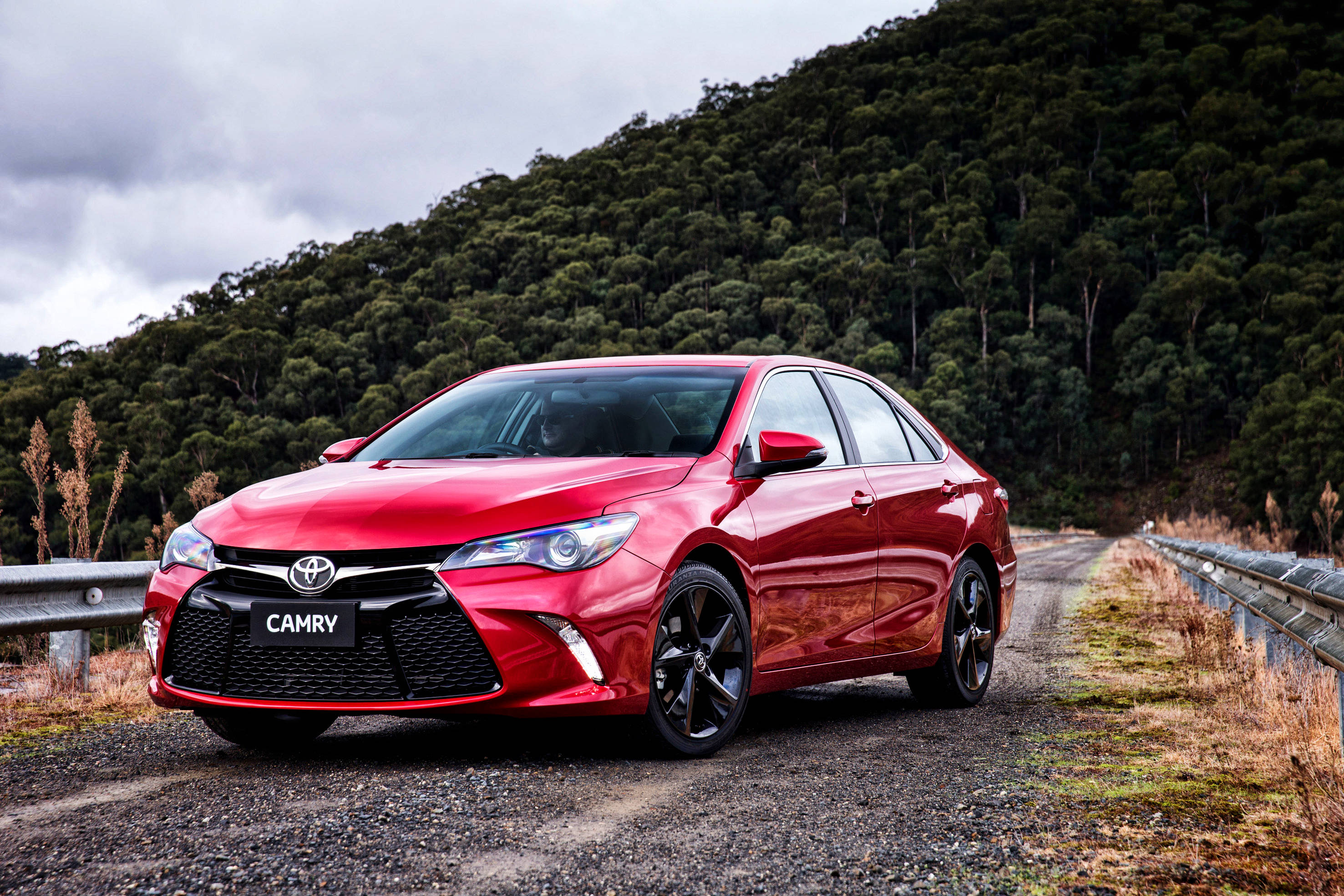 Toyota 桌布 壁紙 — 一輛光澤的紅色豐田Camry轎車停在碎石路上，背景是樹木繁茂的山丘，天空多雲。