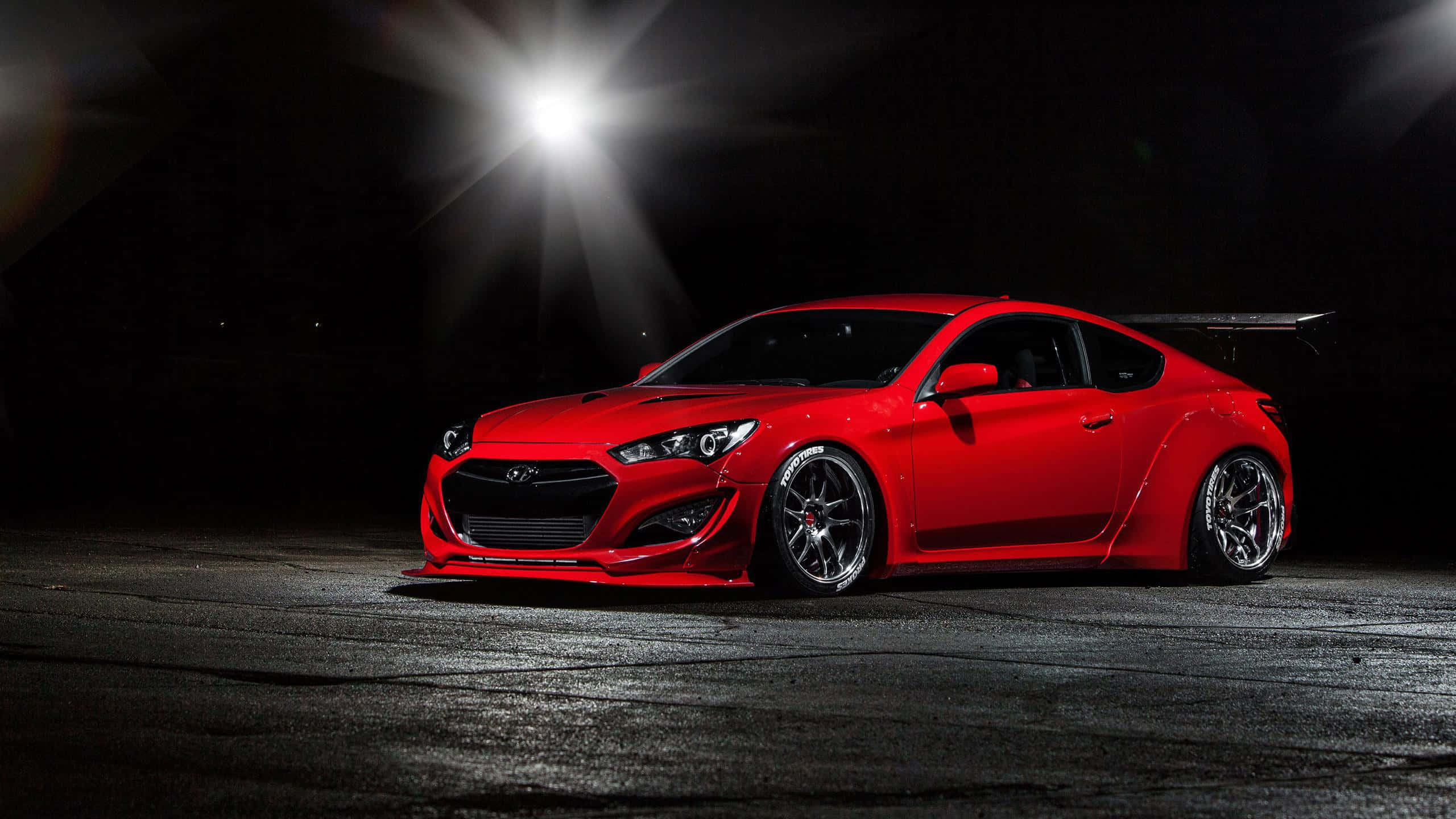 Hyundai Genesis 桌布 壁紙 — 一輛光澤的紅色跑車配備寬體套件和黑色輪圈，在黑暗的瀝青路面上被單一聚光燈戲劇性地照亮。