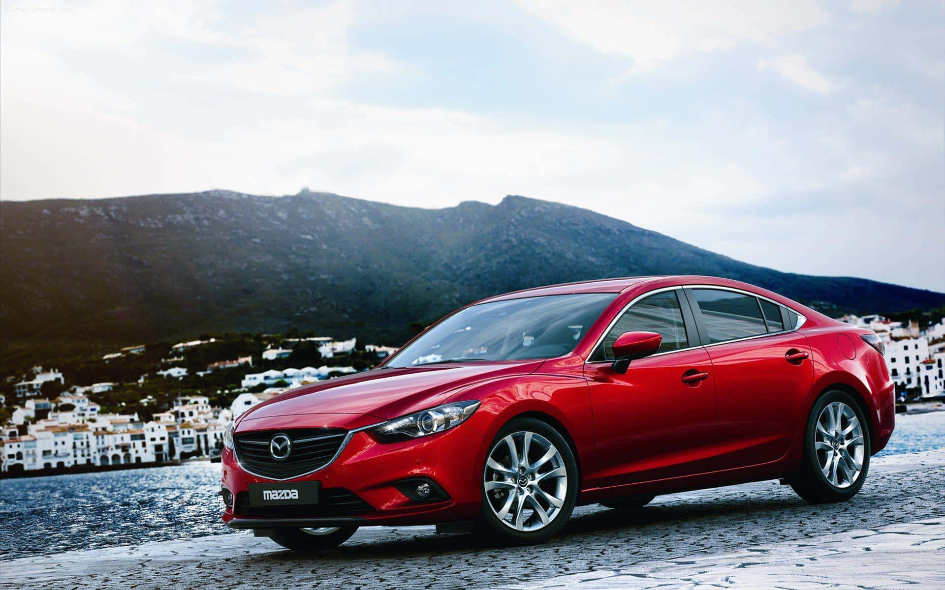 Mazda 6 桌布 壁紙 — 一輛光澤的紅色馬自達房車停泊在鵝卵石海濱，背景有白色建築物和山脈。