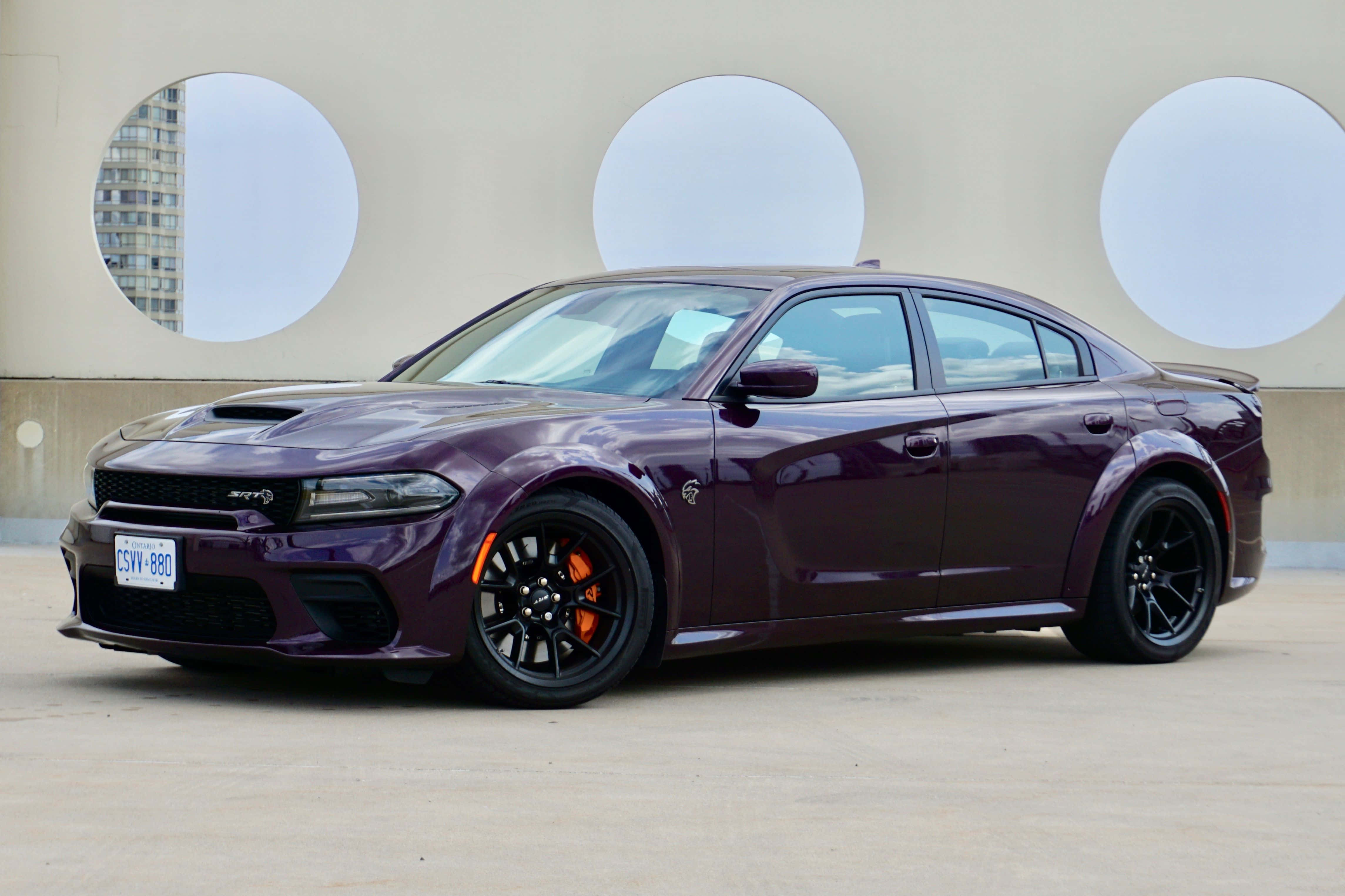 Dodge 桌布 壁紙 — 一輛光澤的紫色 Dodge Charger Hellcat 停在米色牆前，牆上有三個圓形開口，露出城市景觀。