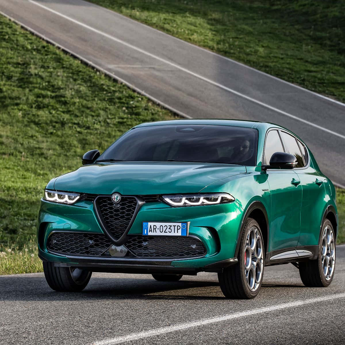 愛快羅密歐 桌布 壁紙 — 一輛光澤的綠色Alfa Romeo SUV在明亮的日光下行駛在一條蜿蜒的瀝青路上，兩旁是茂密的綠草。