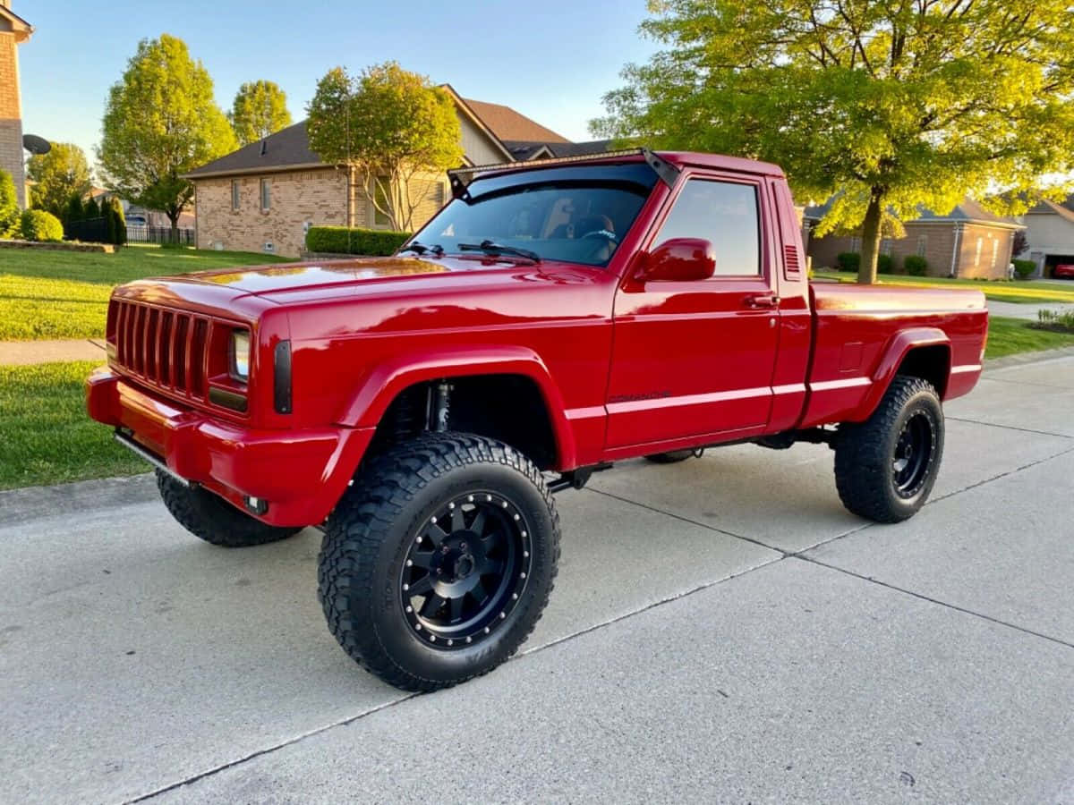 Jeep Comanche 桌布 壁紙 — 一輛光澤紅色升高底盤的Jeep Cherokee皮卡車，配黑色輪胎，在黃金時段停放在郊區社區的混凝土車道上。