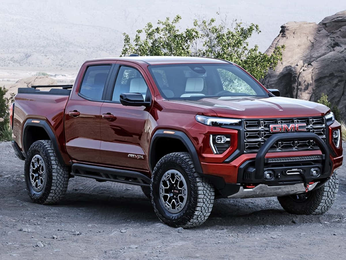 Gmc Canyon 桌布 壁紙 — 一輛光澤紅色的GMC AT4X皮卡車，帶有黑色飾邊和堅固輪胎，停放在岩石地形上，背景是稀疏的綠色植物和大石塊。