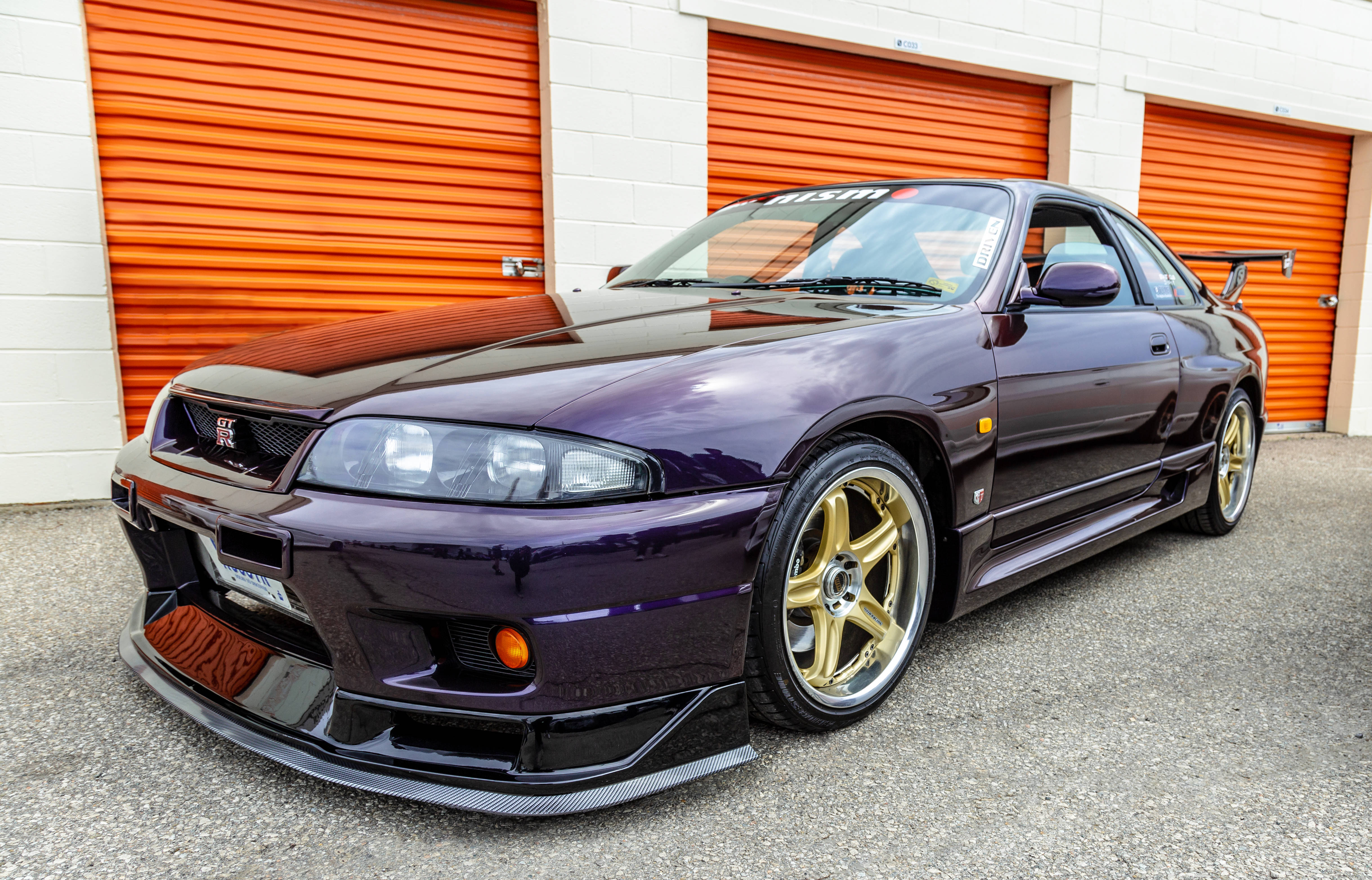 R 桌布 壁紙 — 一輛光澤紫色日產Skyline R34跑車配金色輪圈，停在亮橙色車庫門前。