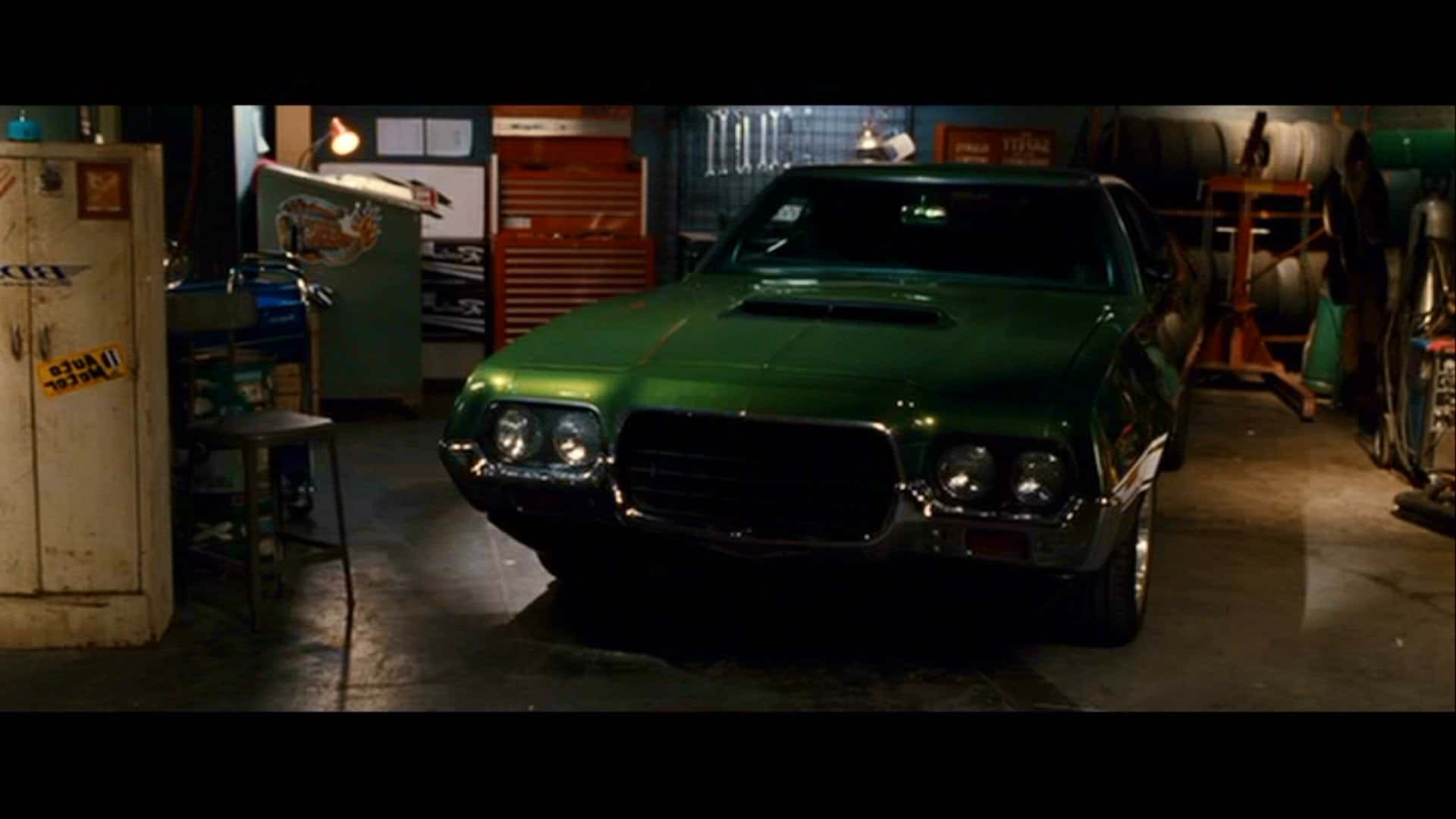 Fast And Furious 1 桌布 壁紙 — 一輛光澤綠色經典肌肉車在雜亂的車庫中，在工場燈光下閃閃發光，車庫裡充滿了工具和設備。