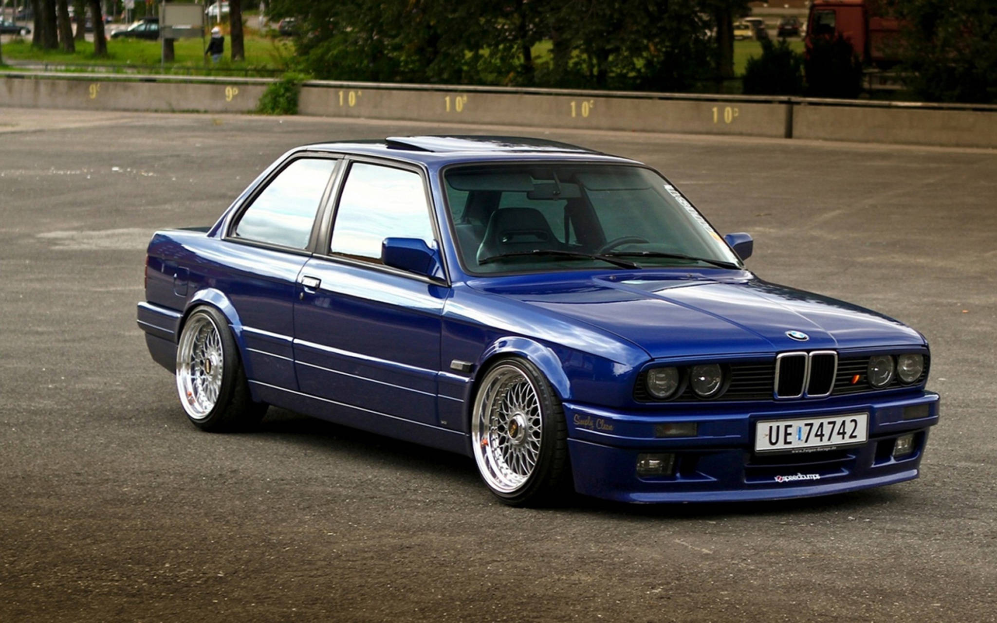 classic bmw 桌布 壁紙 — 一輛光澤藍色BMW E30雙門轎跑車，配備訂製輪圈，停在綠樹環繞的瀝青停車場上。
