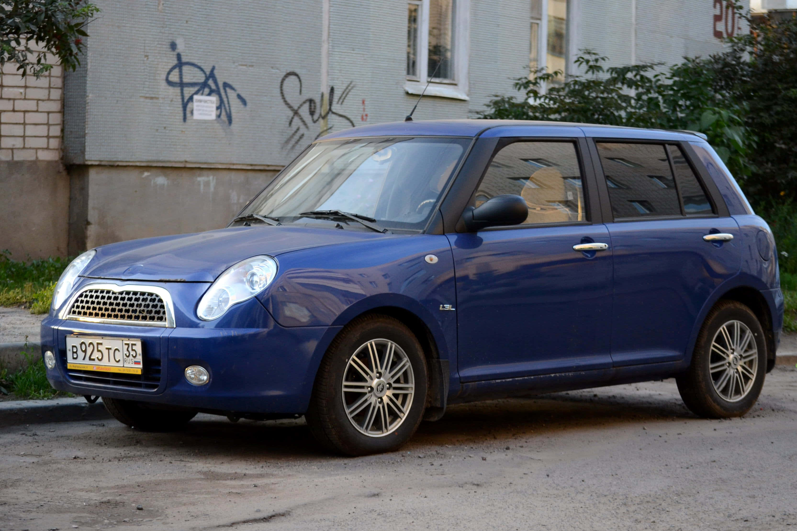 Lifan 桌布 壁紙 — 一輛光澤藍色小型車停泊在裂開的瀝青街道上，旁邊是帶有塗鴉和綠色植物的建築物。