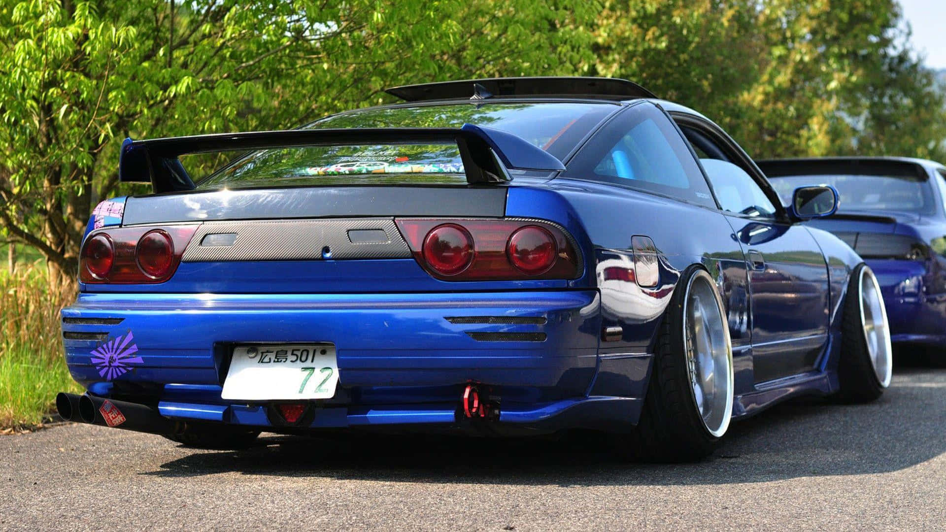 Nissan 180sx 桌布 壁紙 — 一輛光澤藍色日產Skyline R34，配備大型尾翼和定制輪圈，在明亮陽光下停在瀝青路上。