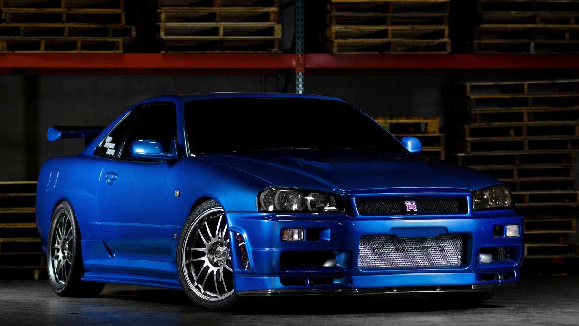 藍色 桌布 壁紙 — 一輛光澤藍色的日產Skyline R34跑車，配備定制輪轂和後擾流板，停在光線昏暗的倉庫中，背景有堆疊的木托盤。