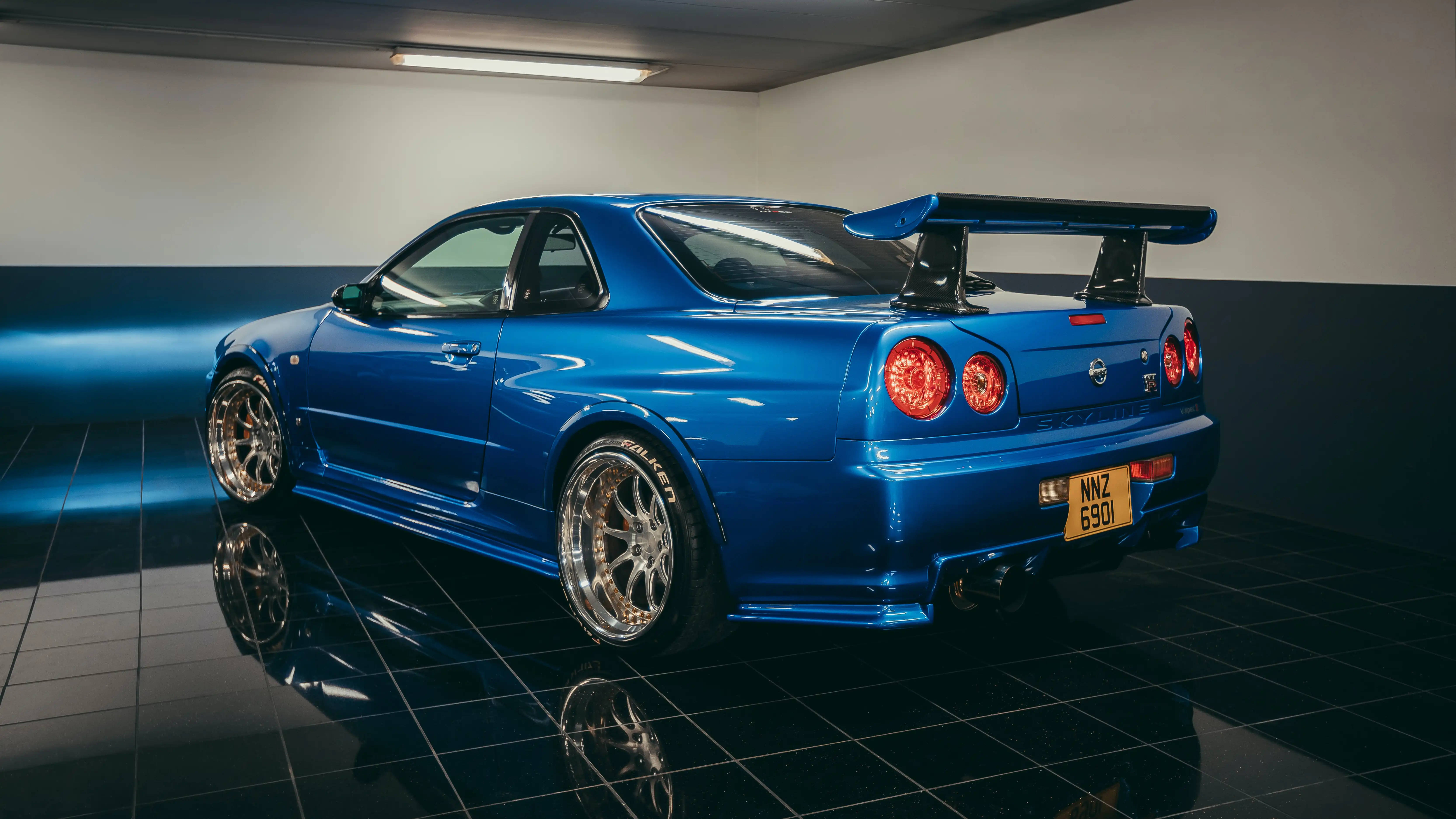 Nissan 桌布 壁紙 — 一輛光澤藍色的日產Skyline R34跑車，帶有大型後擾流板，在光線昏暗、鋪有反光黑色瓷磚地板的車庫中展示。