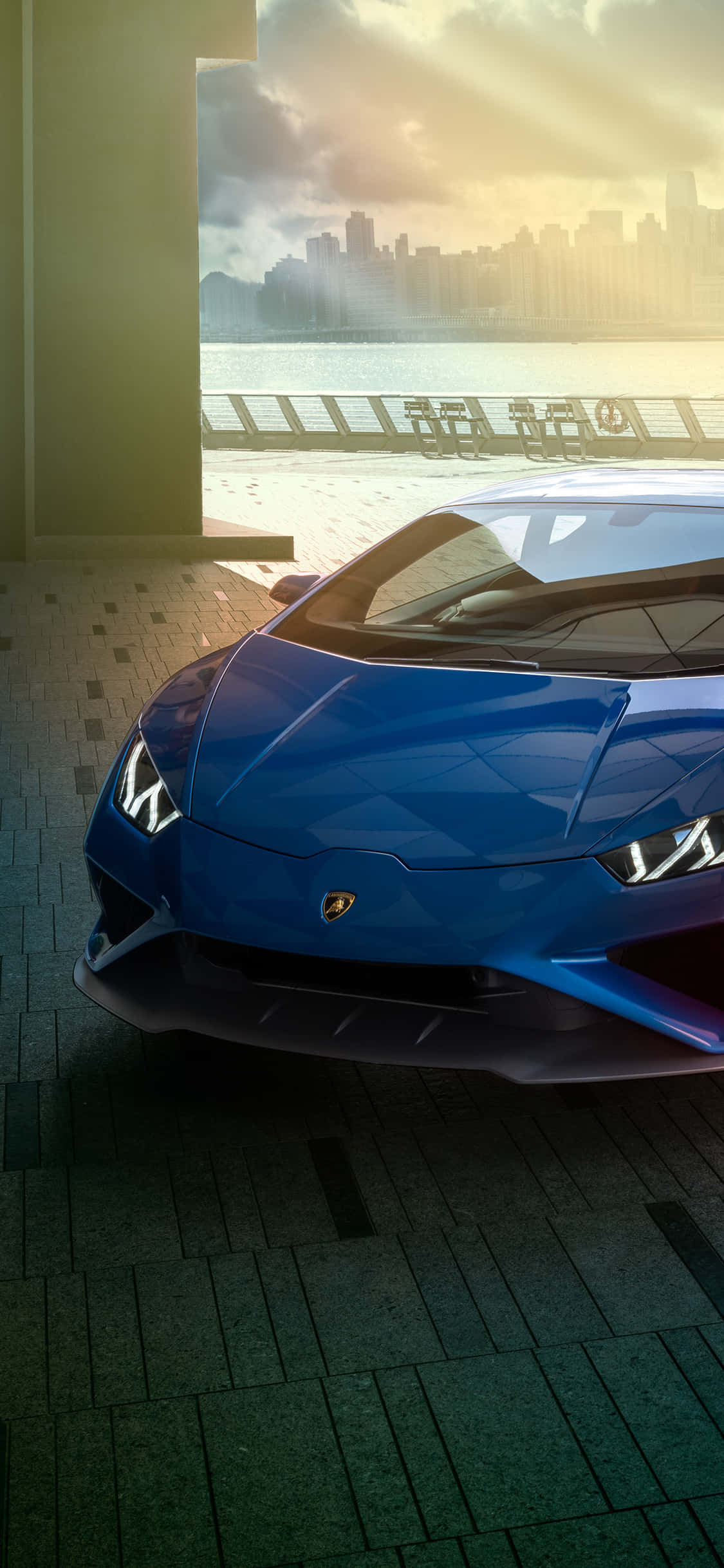 Blue Lamborghini iPhone 桌布 壁紙 — 一輛光澤藍色藍寶堅尼跑車停放在室內，透過大窗戶可以看到夕陽下的城市天際線和水面。