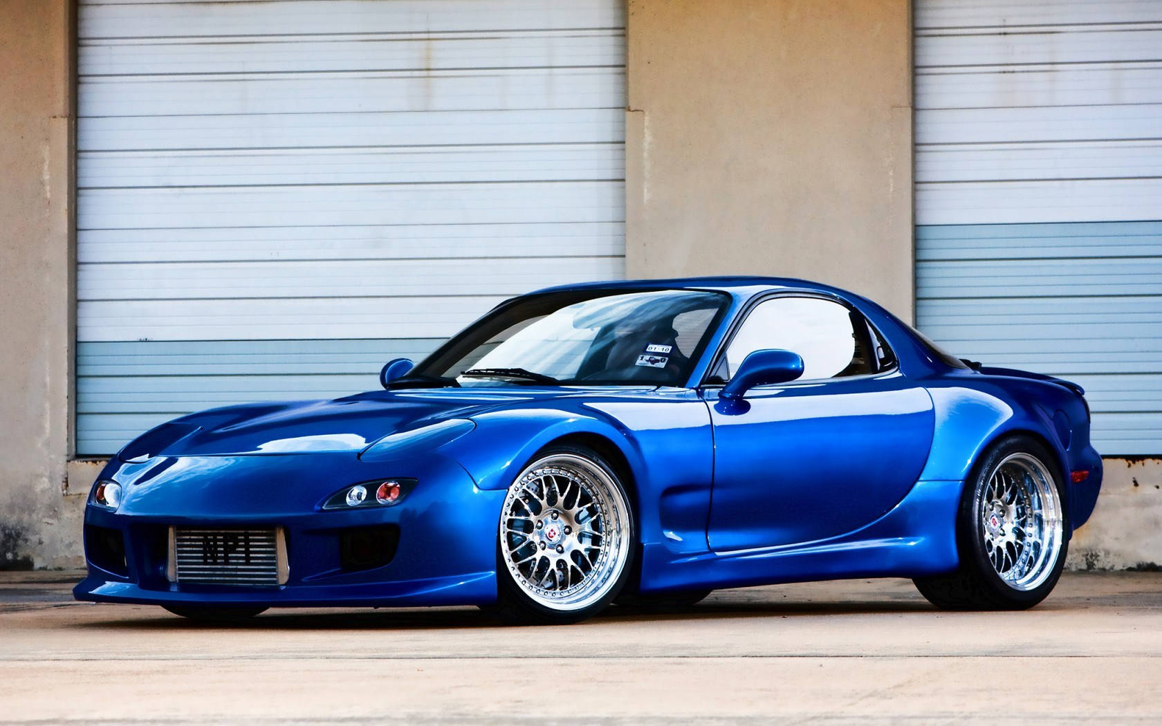Mazda 桌布 壁紙 — 一輛光澤藍色馬自達RX-7跑車配備定制輪圈，停泊在波紋金屬車庫門前。