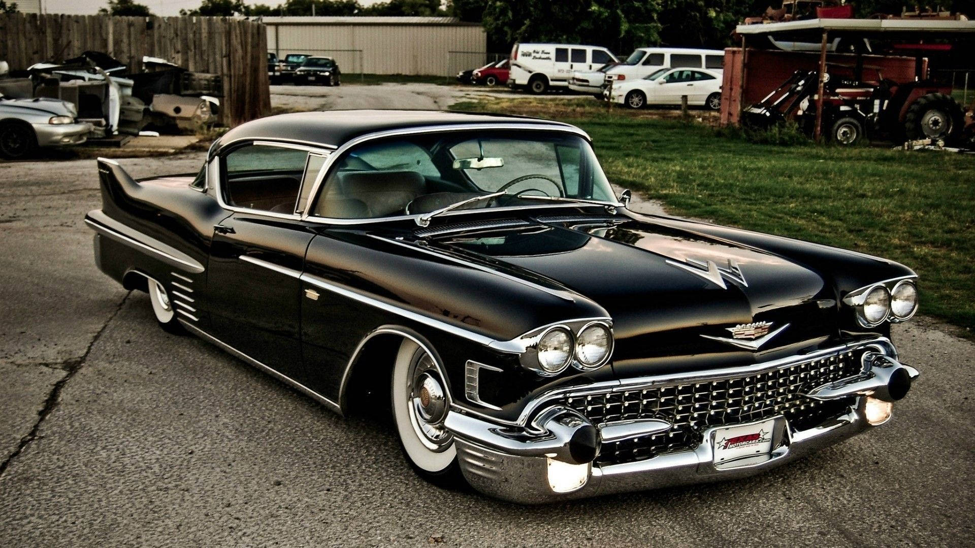 Cadillac 桌布 壁紙 — 一輛光澤黑色1959年凱迪拉克，帶有鍍鉻裝飾和白色輪胎，停在雜亂庭院中破裂的瀝青上。