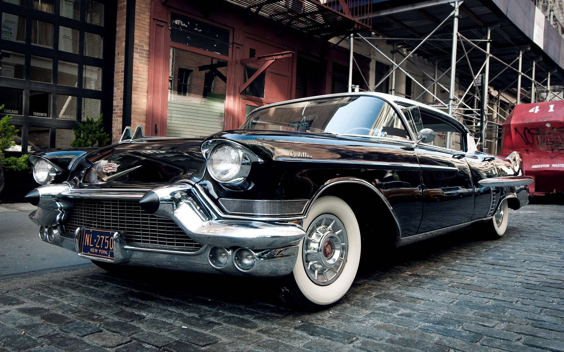Cadillac 桌布 壁紙 — 一輛光澤黑色1959年凱迪拉克，帶有鍍鉻飾邊和白色輪胎，在鵝卵石街道上閃閃發光，旁邊是帶有鷹架的磚砌建築。