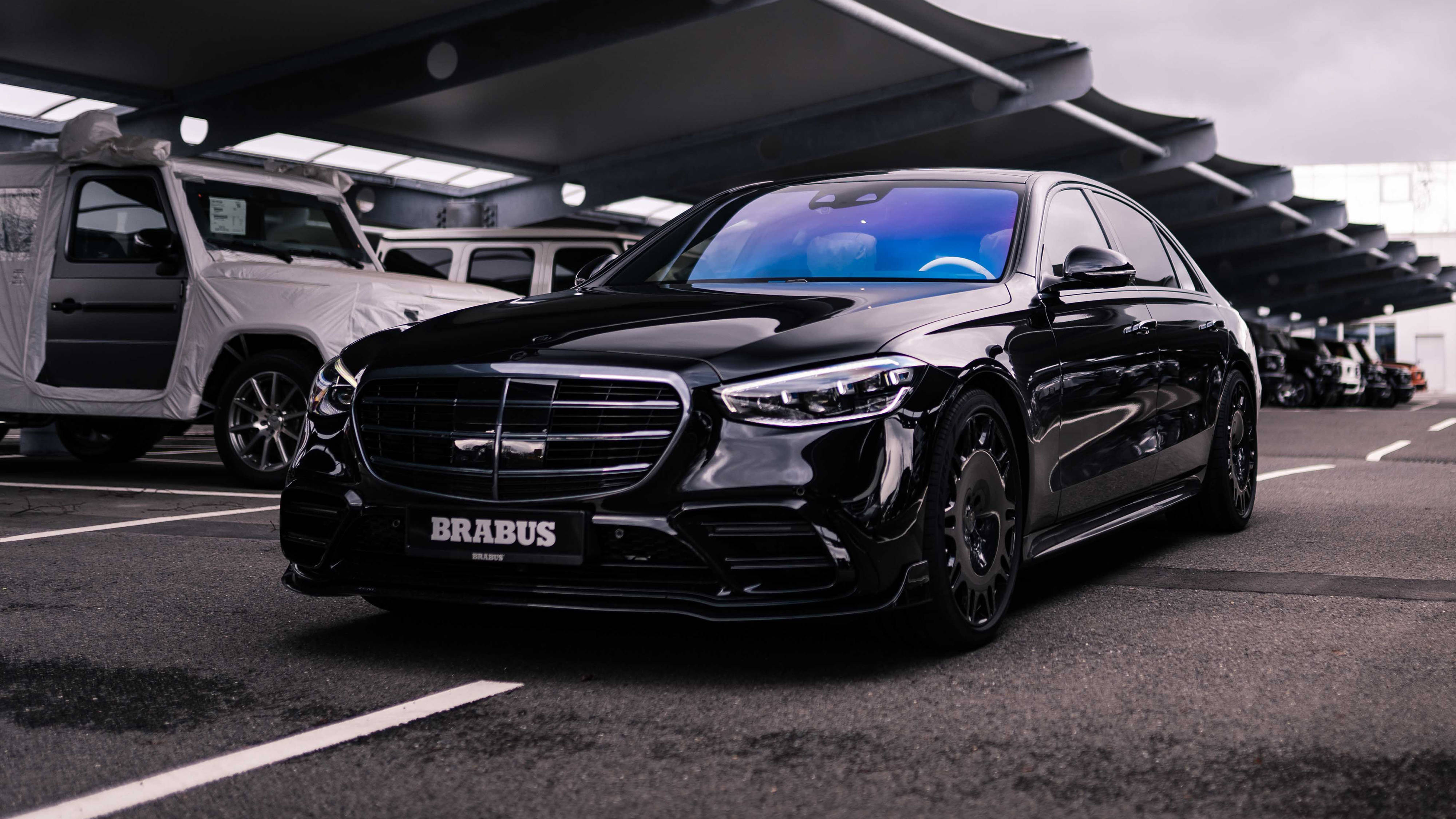 4K 奔馳壁紙 桌布 壁紙 — 一輛光澤黑色Brabus Mercedes轎車在陰天下的現代有蓋停車場中閃耀。