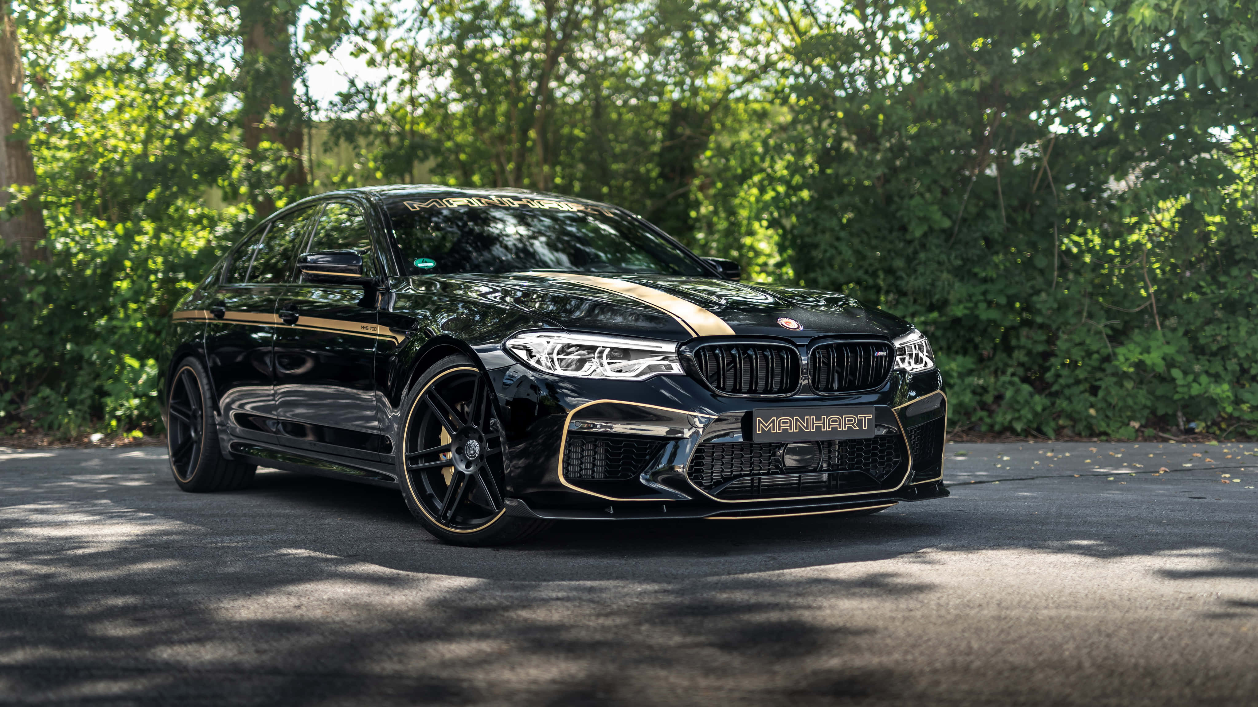 桌布 壁紙 — 一輛光澤黑色Manhart改裝BMW M5，帶有金色點綴，停在綠樹成蔭的柏油路上。