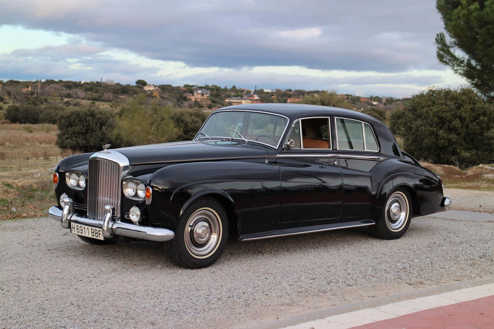 Bentley S3 桌布 壁紙 — 一輛光澤黑色復古賓利轎車停在碎石上，背後是丘陵景觀和多雲的天空。