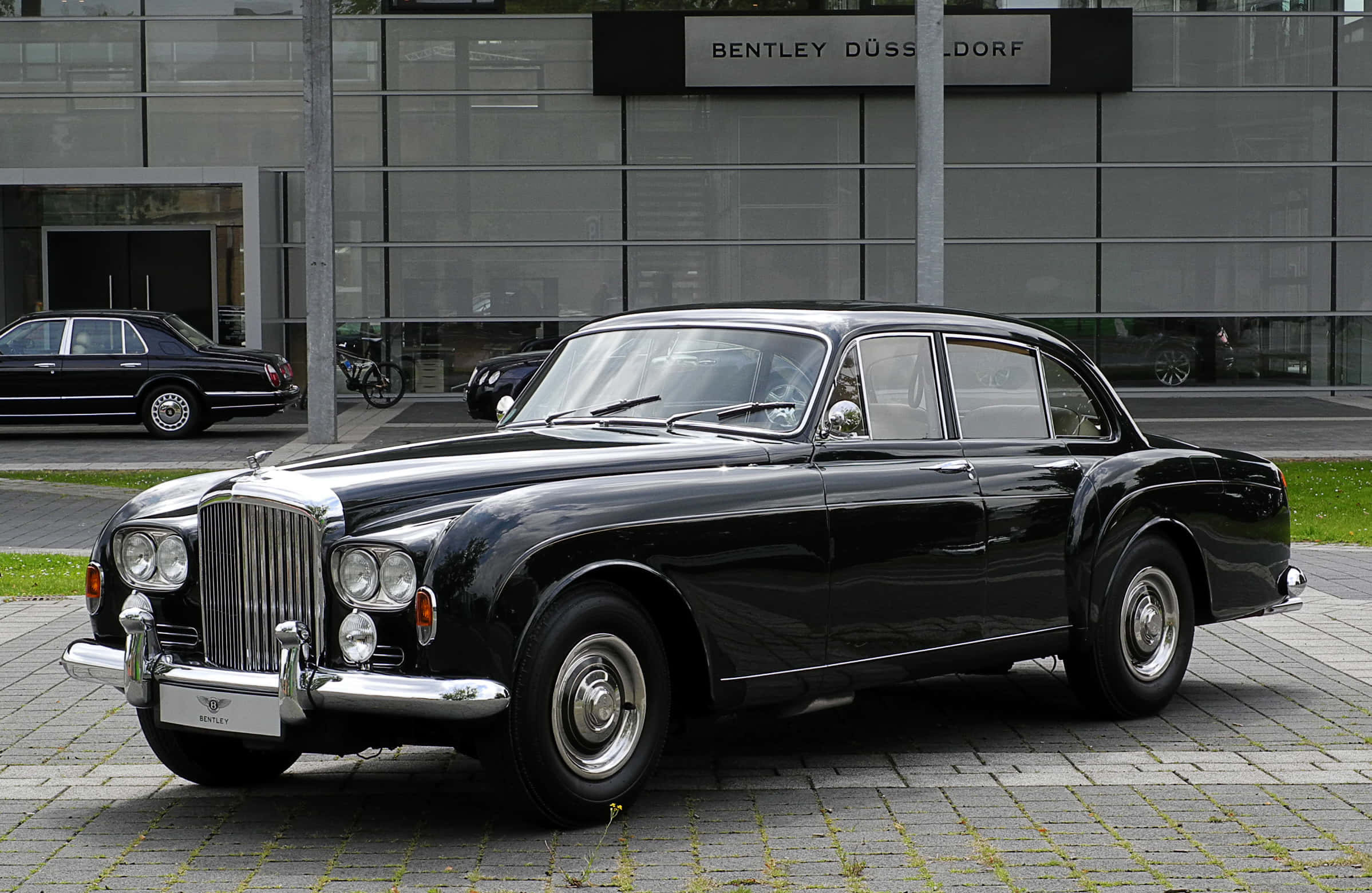 Bentley S3 桌布 壁紙 — 一輛光澤黑色復古賓利轎車停放在現代玻璃建築前的鋪砌庭院。