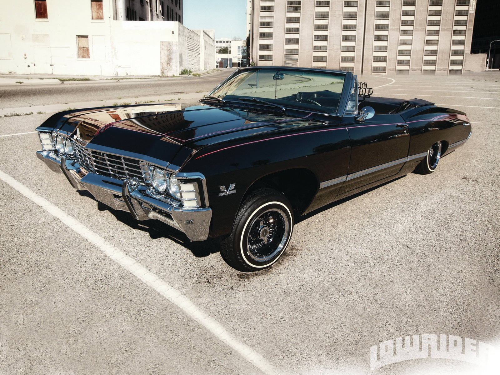 Lowrider 桌布 壁紙 — 一輛光澤黑色經典敞篷車，帶有鍍鉻裝飾，停在空曠的城市地塊上，在明亮的日光下。