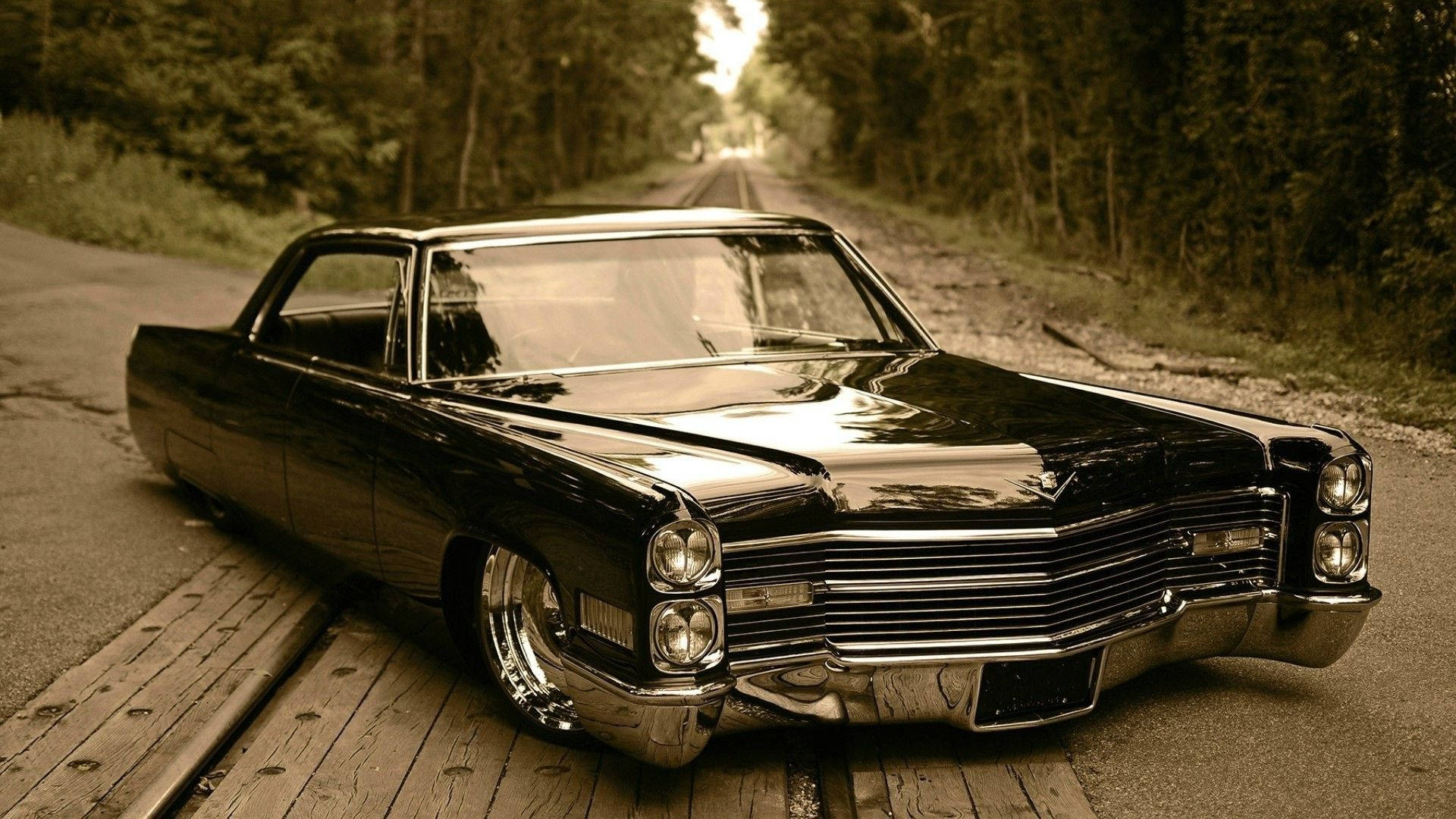 Cadillac 桌布 壁紙 — 一輛光澤黑色經典汽車帶有鍍鉻細節，停在木板上，旁邊是樹木成蔭的道路，在溫暖的棕褐色光線下。