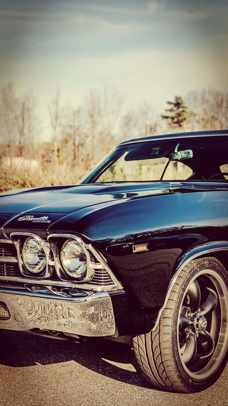 Vintage Car iPhone 桌布 壁紙 — 一輛光澤黑色經典肌肉車，帶有鍍鉻細節，停在瀝青路面上，背景是蒼白的天空和光禿禿的樹木。