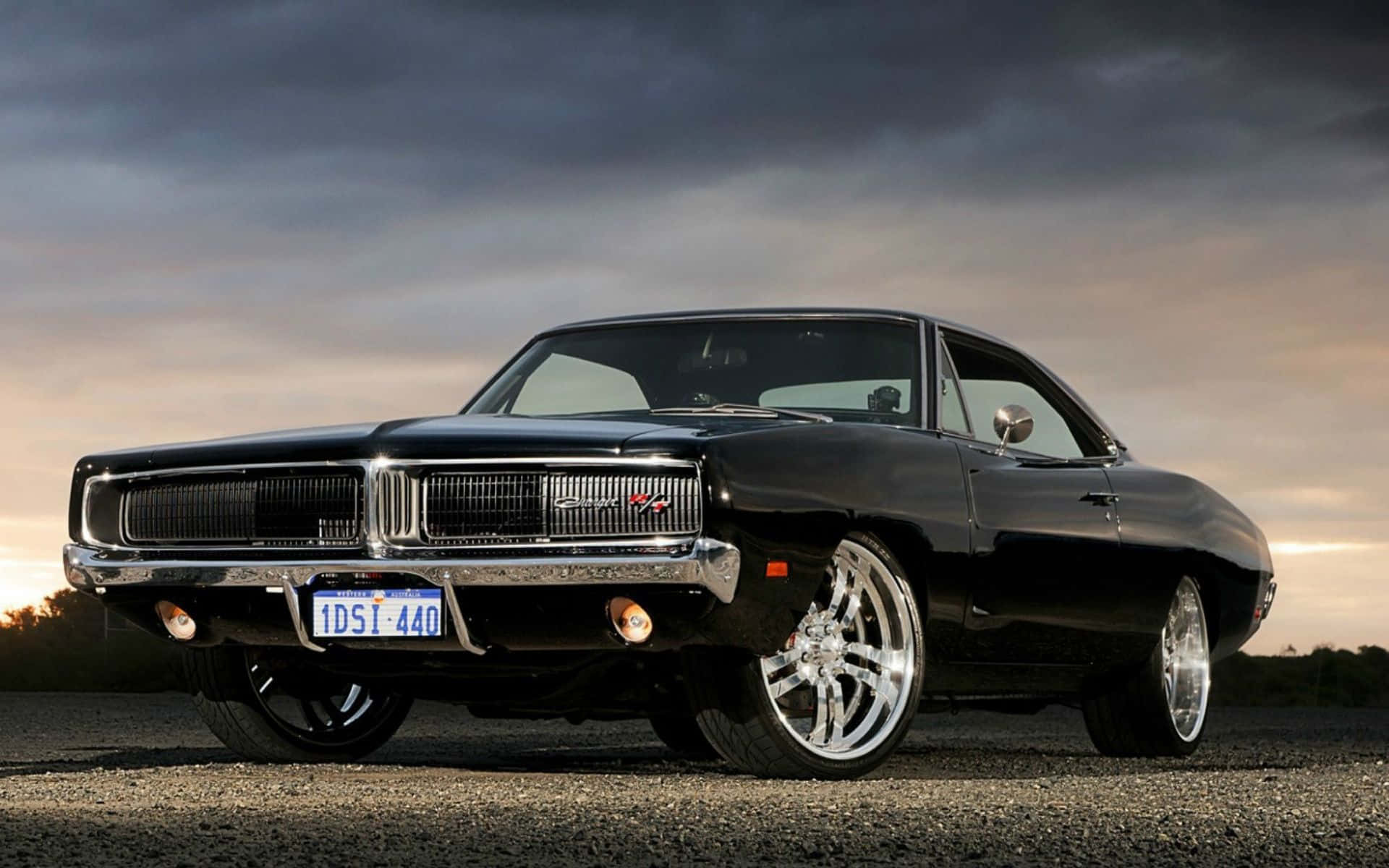 Dodge Charger 桌布 壁紙 — 一輛光澤黑色經典肌肉車，帶有鍍鉻裝飾和訂製輪圈，停在碎石地上，背景是戲劇性的暮色天空。