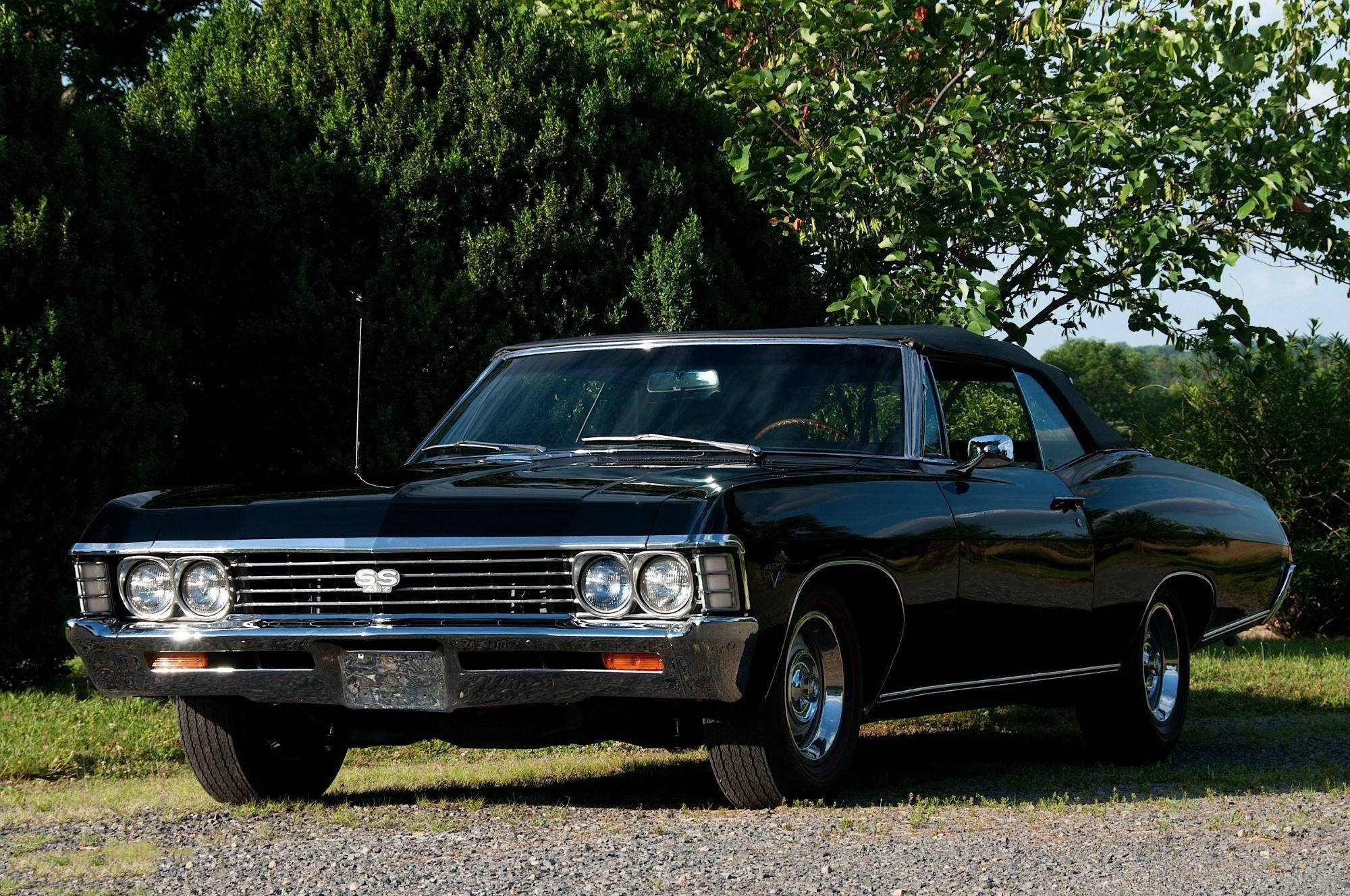 chevrolet impala 1967 桌布 壁紙 — 一輛光澤黑色經典肌肉車，帶有鍍鉻裝飾，在陽光明媚的日子停泊在礫石上，周圍是枝葉茂密的樹木。