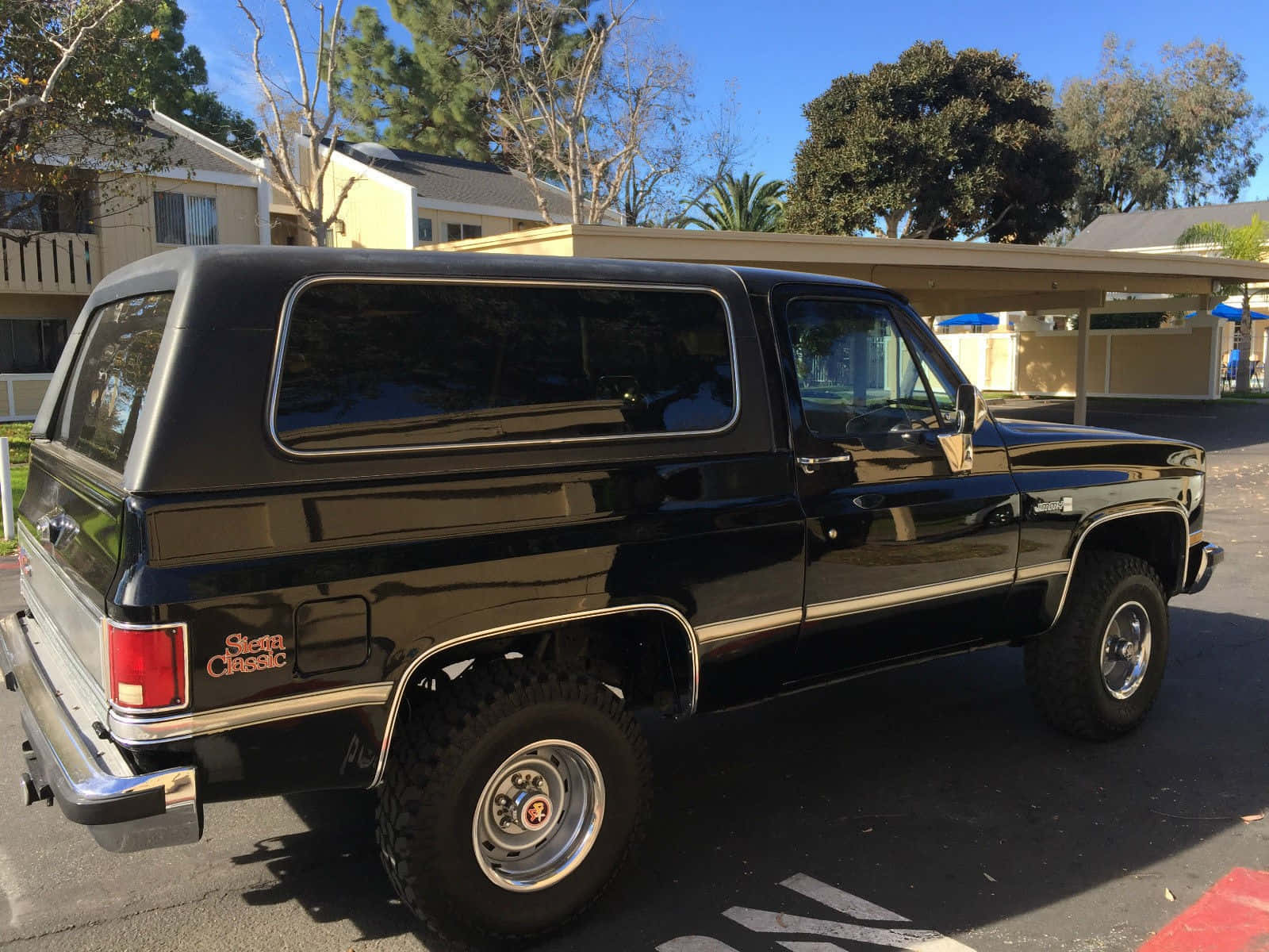 Gmc Jimmy 桌布 壁紙 — 一輛光澤黑色經典雪佛蘭 Blazer SUV 停放在陽光明媚的郊區停車場，背景有棕櫚樹和房屋。
