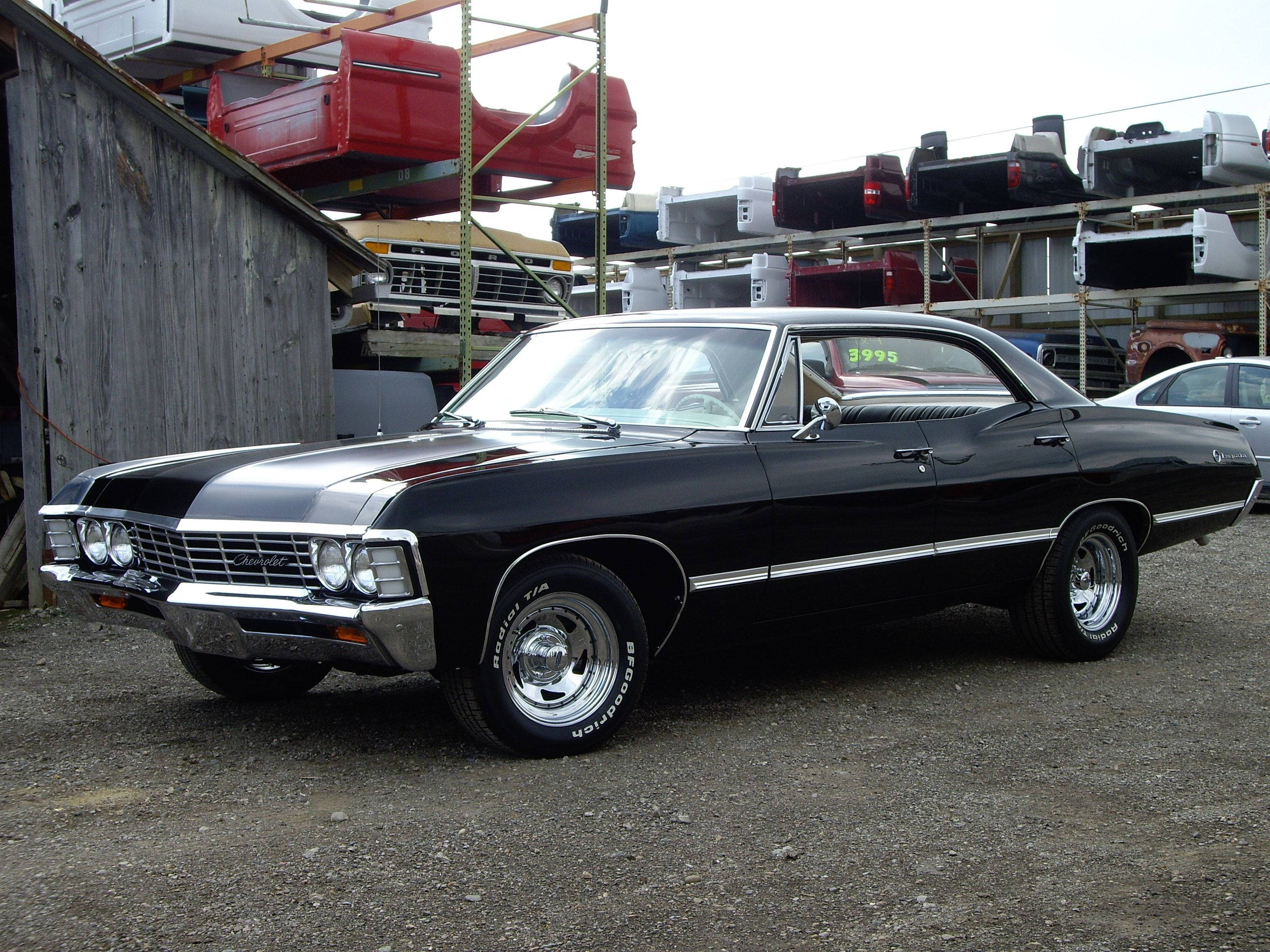 chevrolet impala 1967 桌布 壁紙 — 一輛光澤黑色經典雪佛蘭，帶有鍍鉻裝飾，停放在堆滿汽車零件的倉庫前的碎石地上。