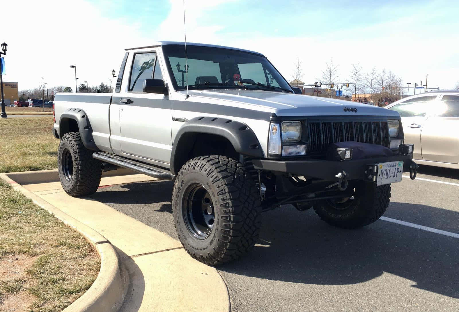 Jeep Comanche 桌布 壁紙 — 一輛升高了的銀色Jeep Cherokee皮卡車，配備超大輪胎，在陽光明媚的日子停放在鋪砌的停車場。