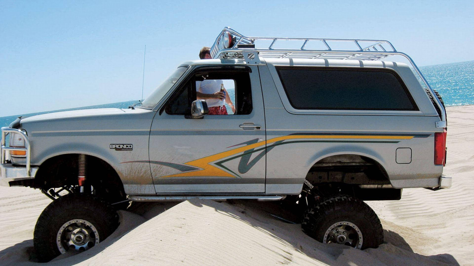 Ford Bronco 桌布 壁紙 — 一輛升高了的銀色福特Bronco帶有黃色條紋，在清澈藍天下沿著碧綠海洋旁的金色沙丘行駛。