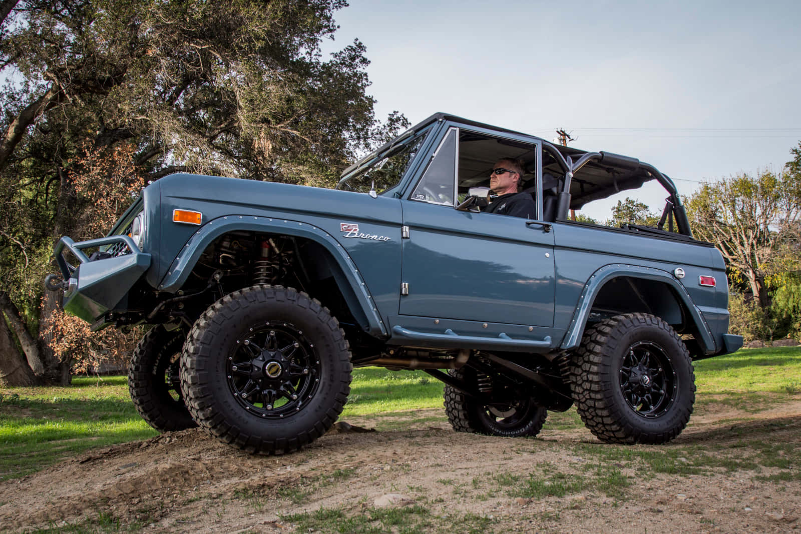 Ford Bronco 桌布 壁紙 — 一輛升高的藍色Bronco配備超大輪胎，停在一個被茂密樹木環繞的泥土路上，天空晴朗。