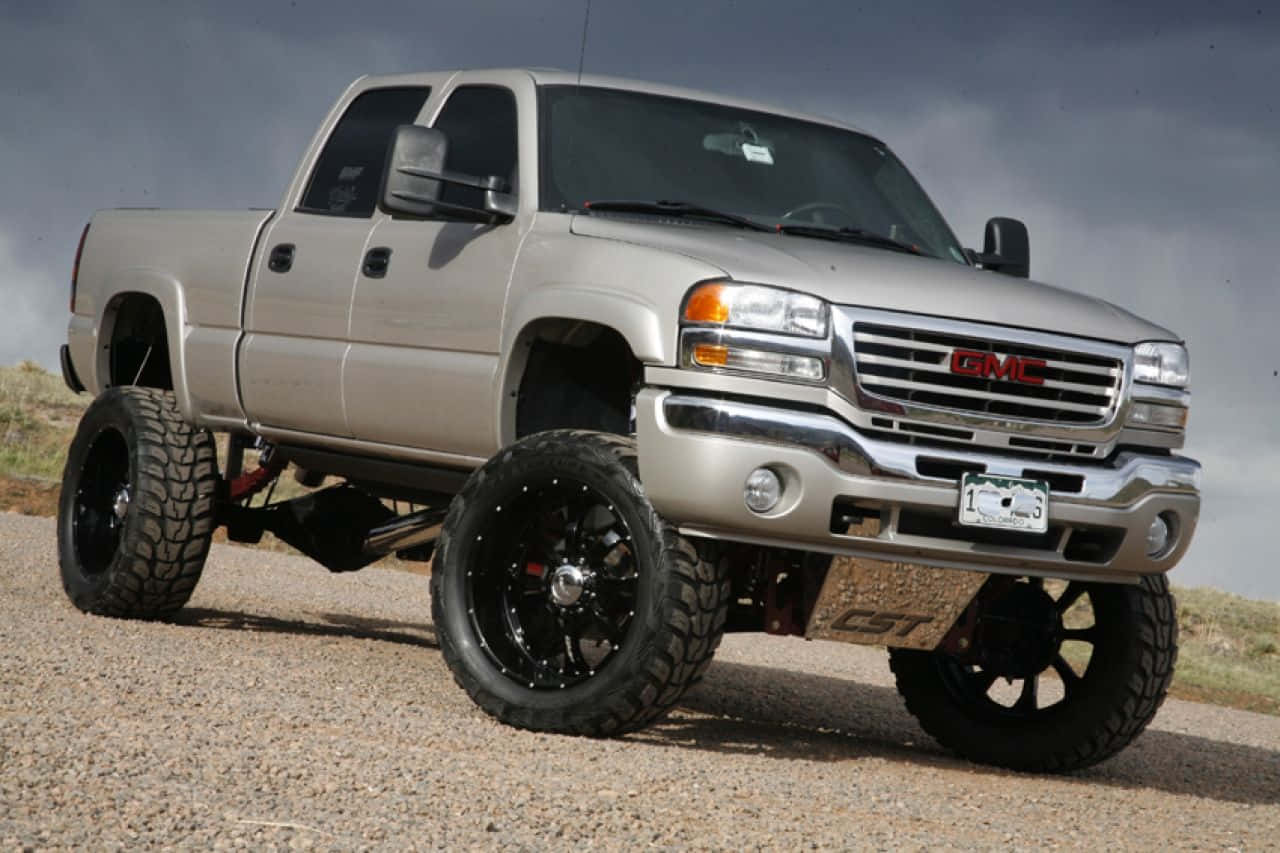 Jacked Up Trucks 桌布 壁紙 — 一輛升高的銀色 GMC Sierra 皮卡車，配備黑色輪圈，停在礫石路上，天空多雲。