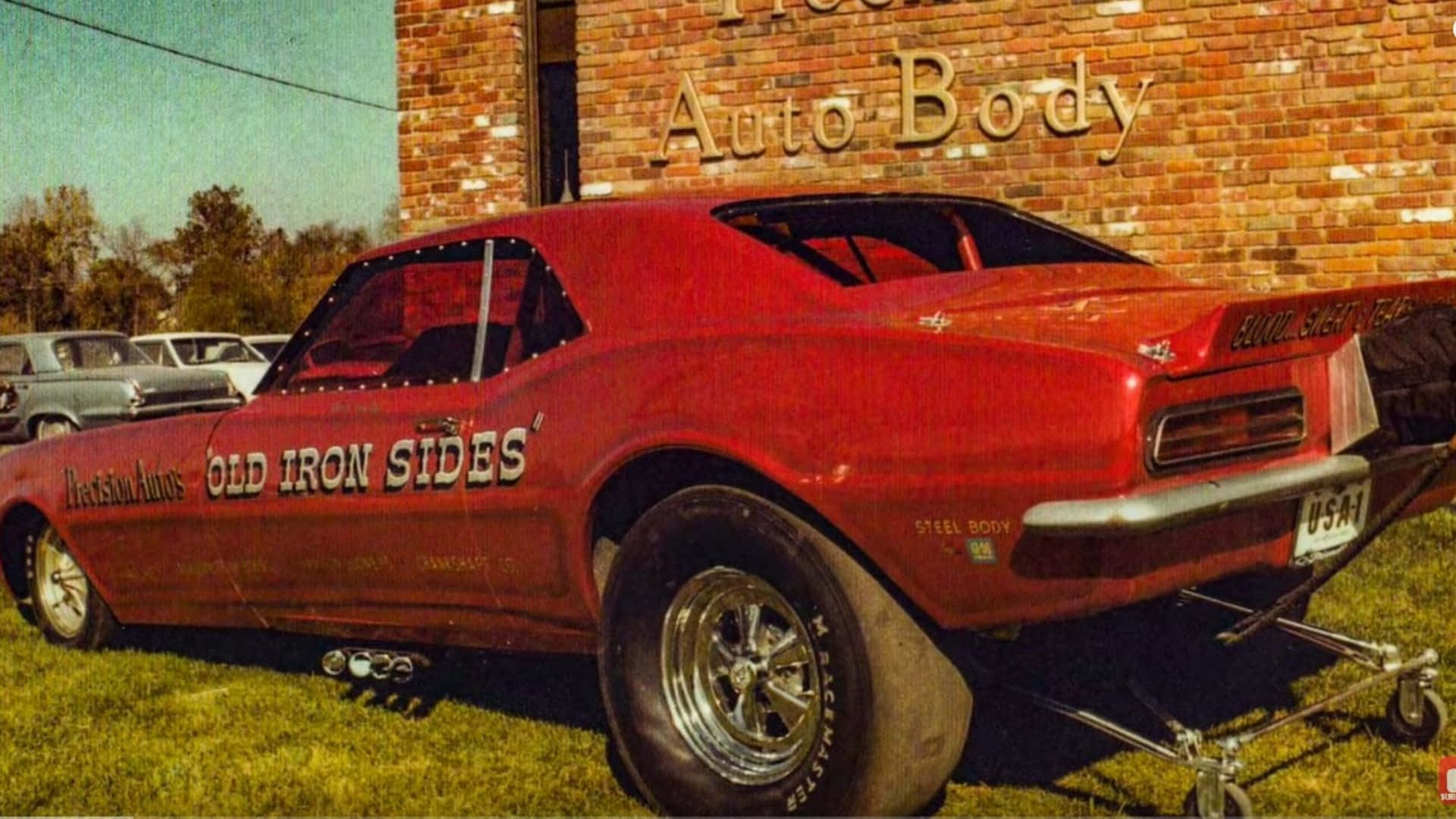 cars 桌布 壁紙 — 一輛名為「Old Iron Sides」的復古紅色直線加速賽車停放在磚造車身修理廠前的草地上，天空清澈湛藍。