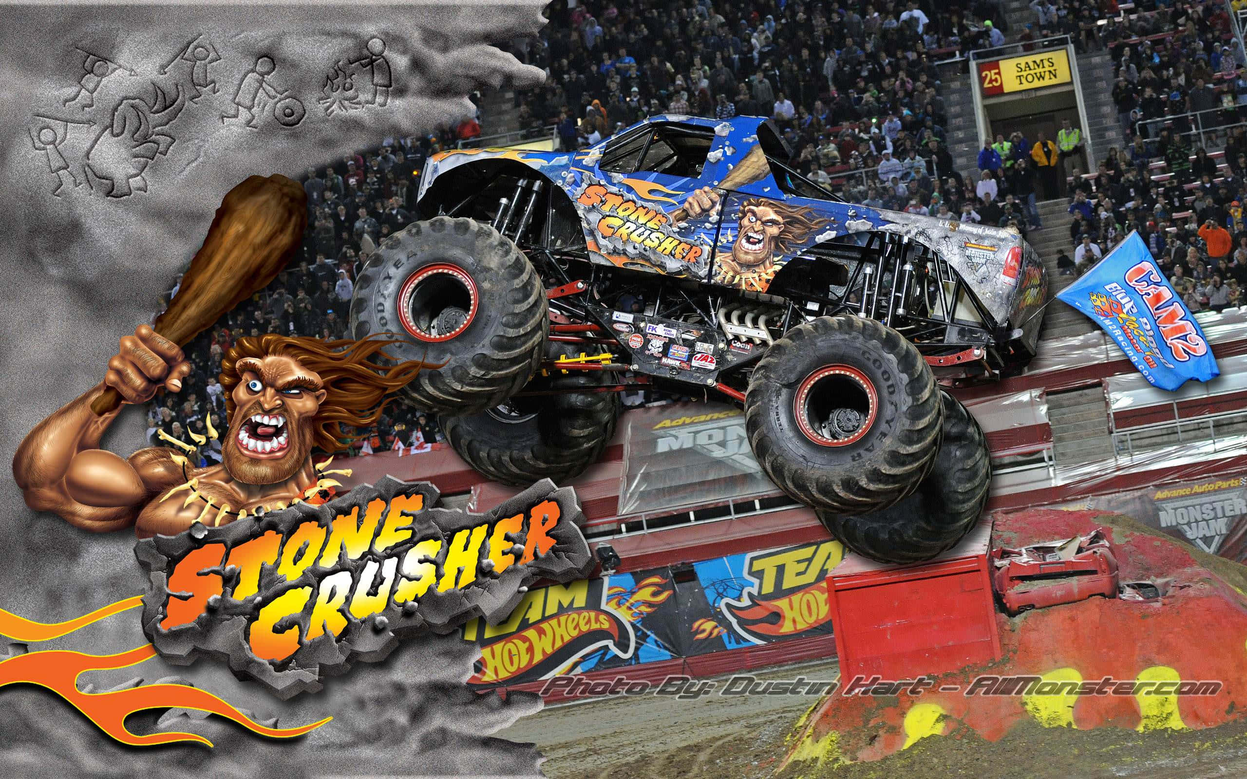 Monster Truck 桌布 壁紙 — 一輛名為 Stone Crusher 的藍色怪獸卡車飛越一輛被壓碎的汽車，同時一個肌肉發達的吉祥物揮舞著球棒，背景是擠滿人群的體育場。